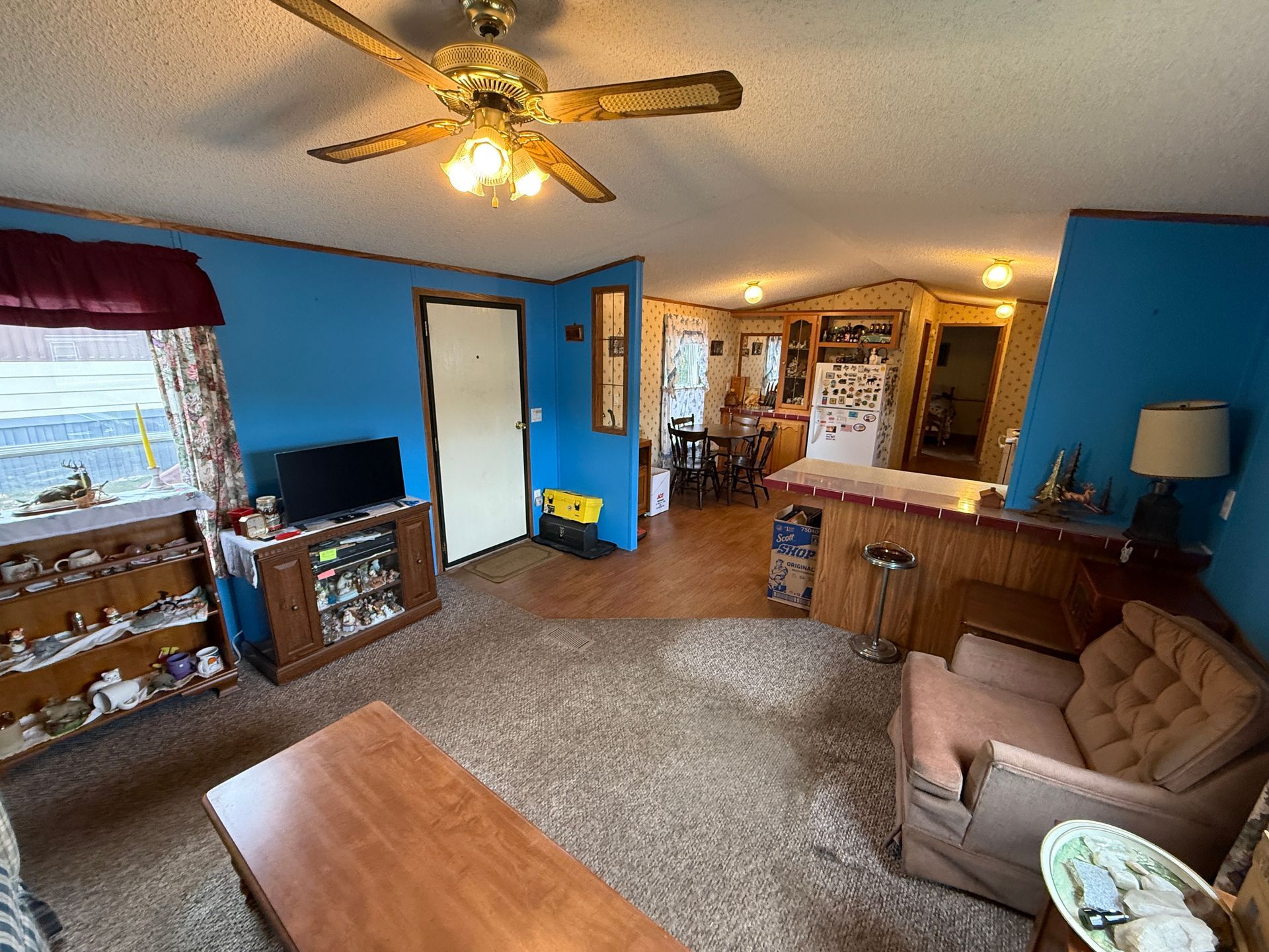 Blue living room with couch, TV, wood table, carpet, and kitchen opening in background
