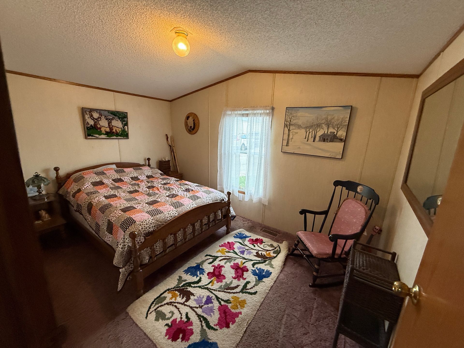 Small bedroom with a bed, floral rug, window, and dresser; warm light and pink chair in the corner.