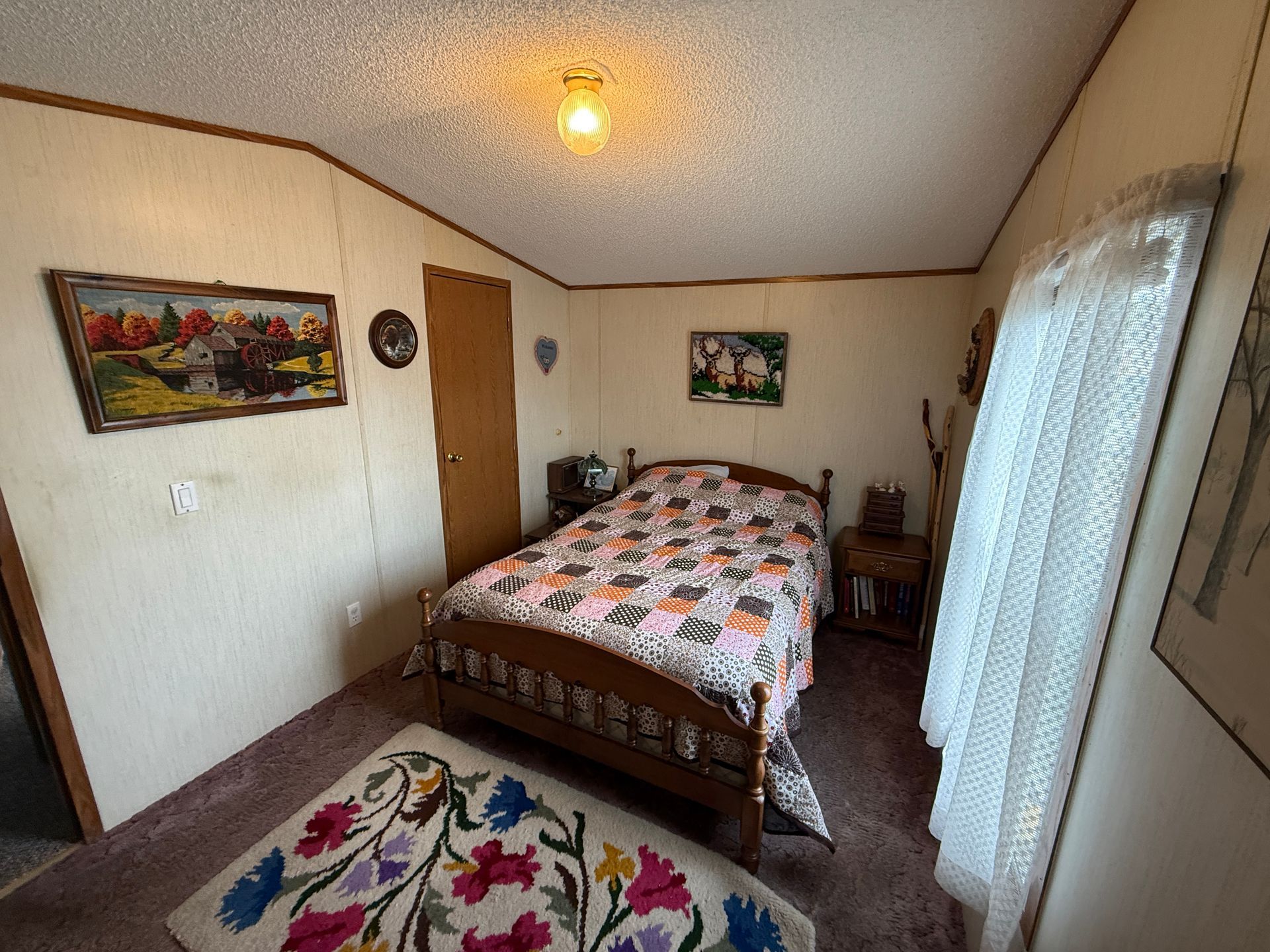 Small bedroom with a neatly made bed, rug, wall art, and lace curtains by a window.