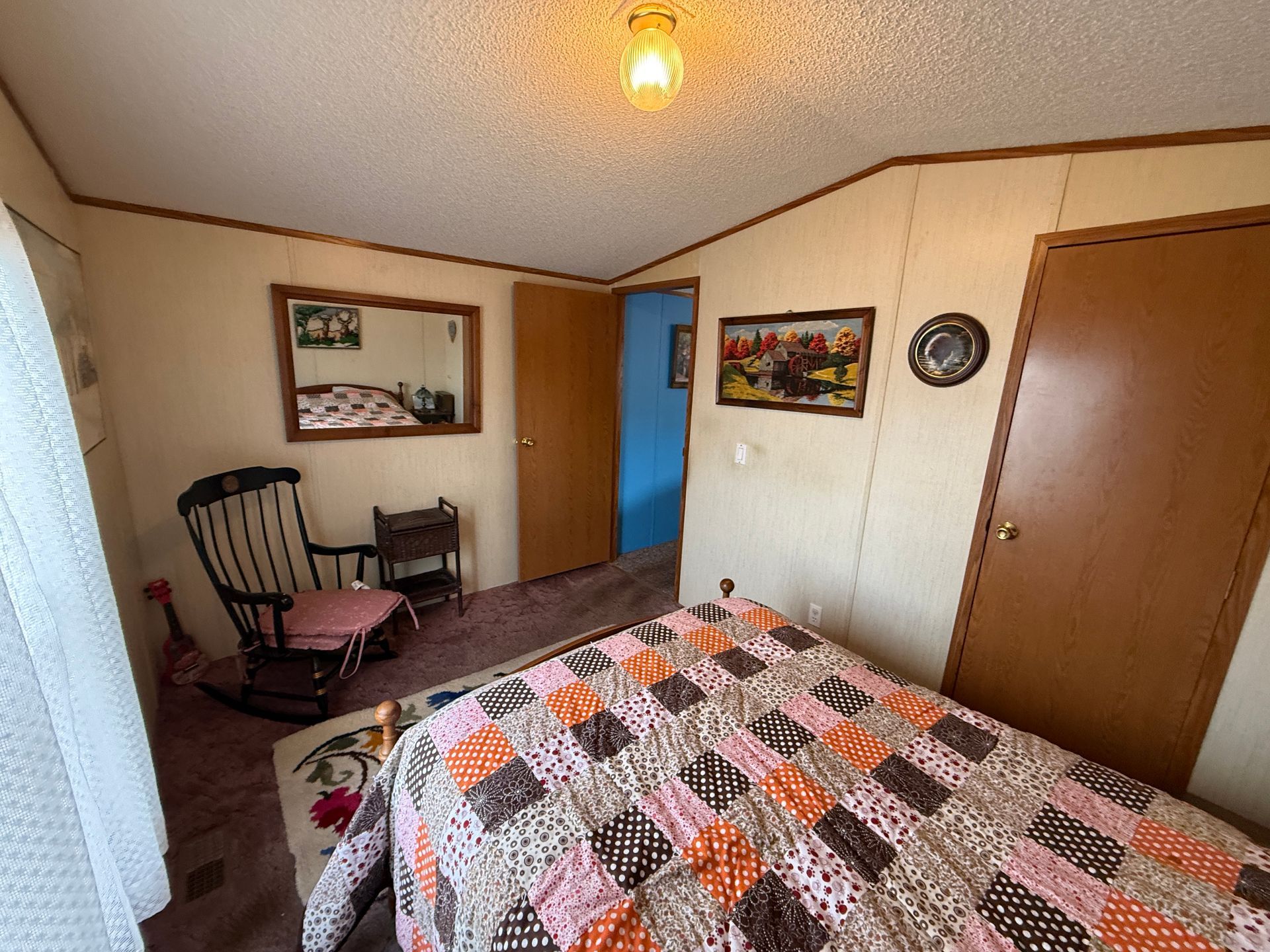 Small bedroom with a quilted bedspread, rocking chair, wall art, and an open door to a blue hallway.