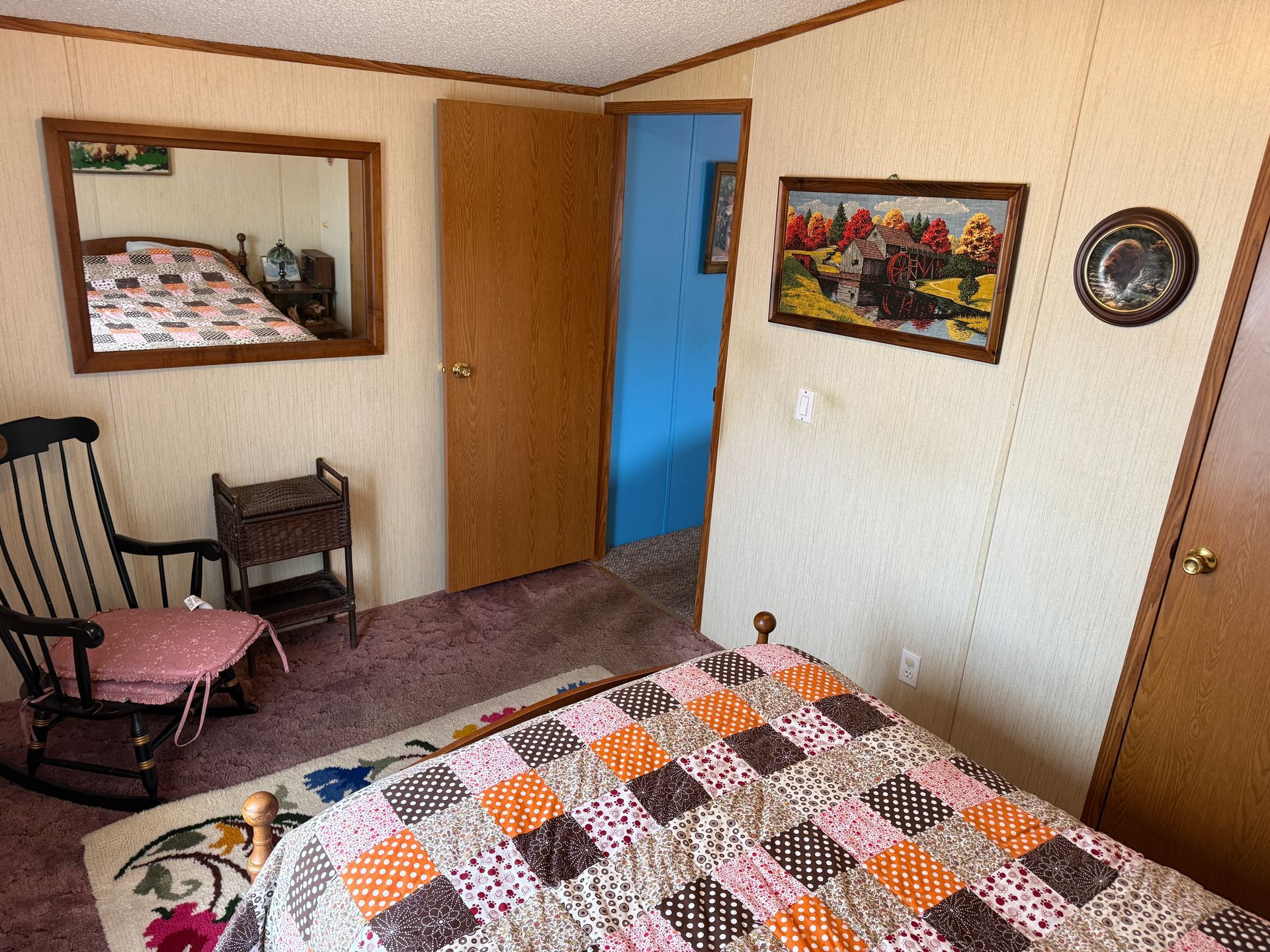 Small bedroom with quilted bed, chair, mirror, wood doors, and framed wall art.