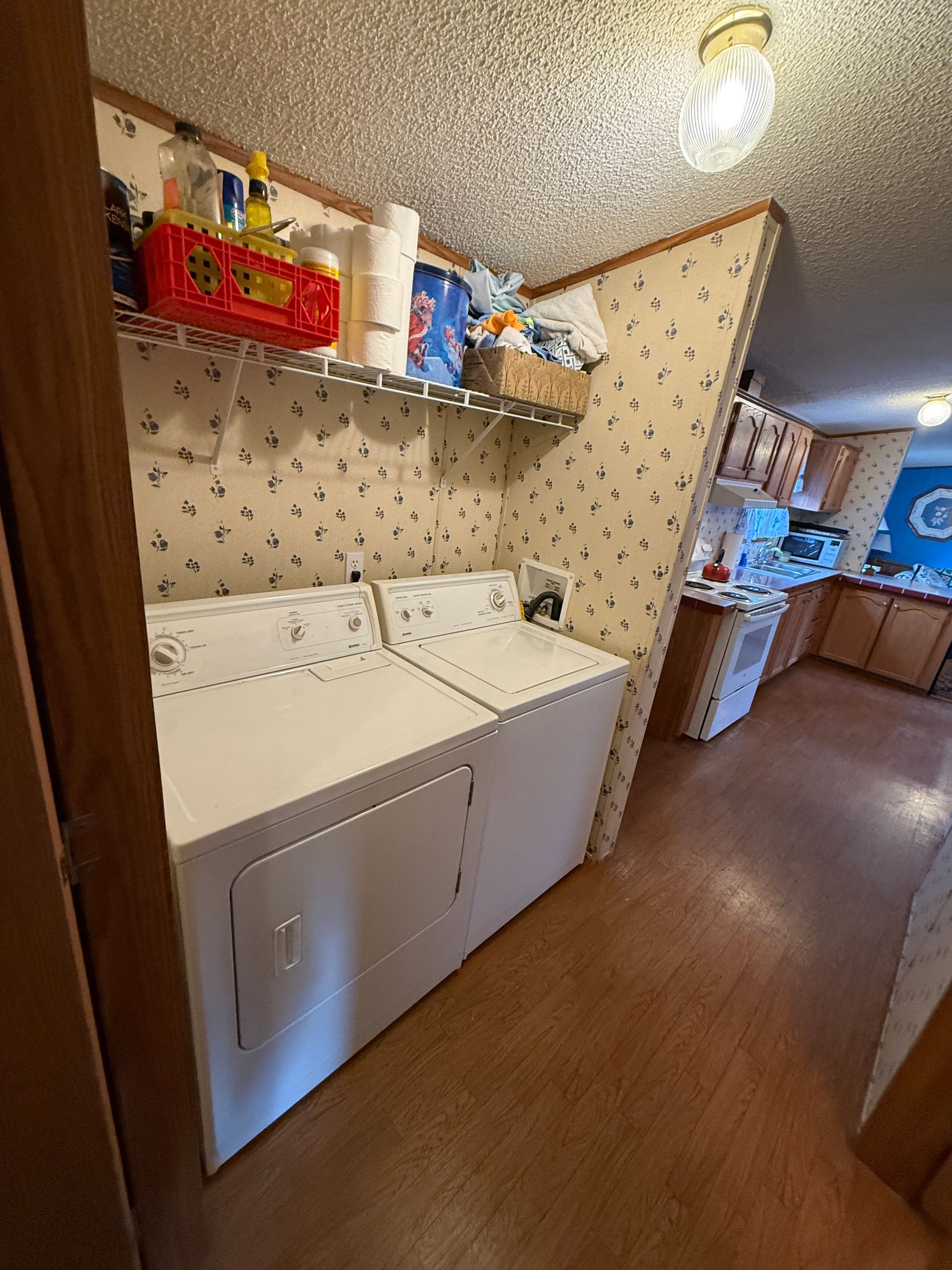 Laundry room with washer and dryer, shelves of detergent, and kitchen area visible in the background.