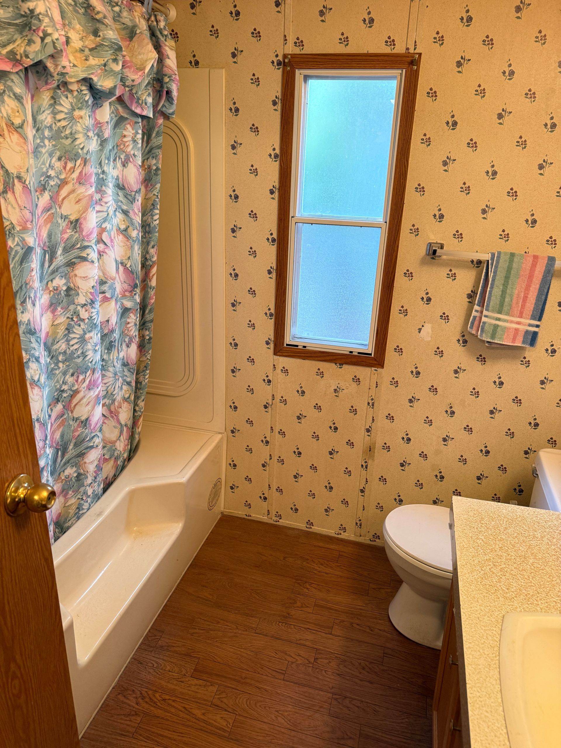 Small bathroom with floral wallpaper, bathtub, toilet, window, and striped towel.