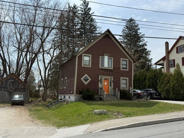 A two-story, brown wooden house with an orange front door, a diamond-shaped window, and surrounding trees on a sunny day.