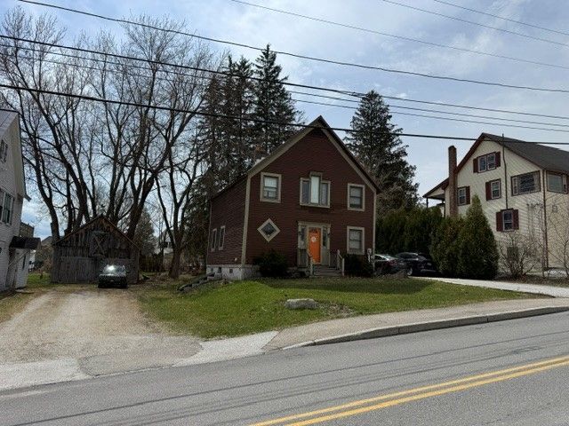 A brown house with a bright orange door stands between a gravel driveway and a light-colored house on a suburban street.