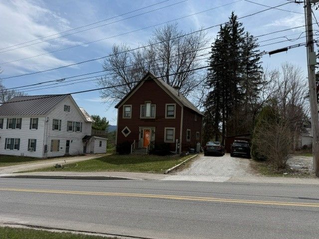 A two-story brown brick house sits along a paved road under a bright, partly cloudy sky.