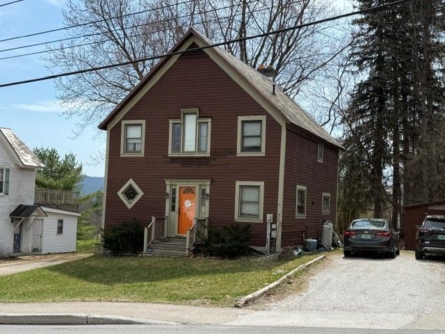 A two-story, reddish-brown house with tan trim, a bright orange front door, and a gravel driveway on a sunny day.