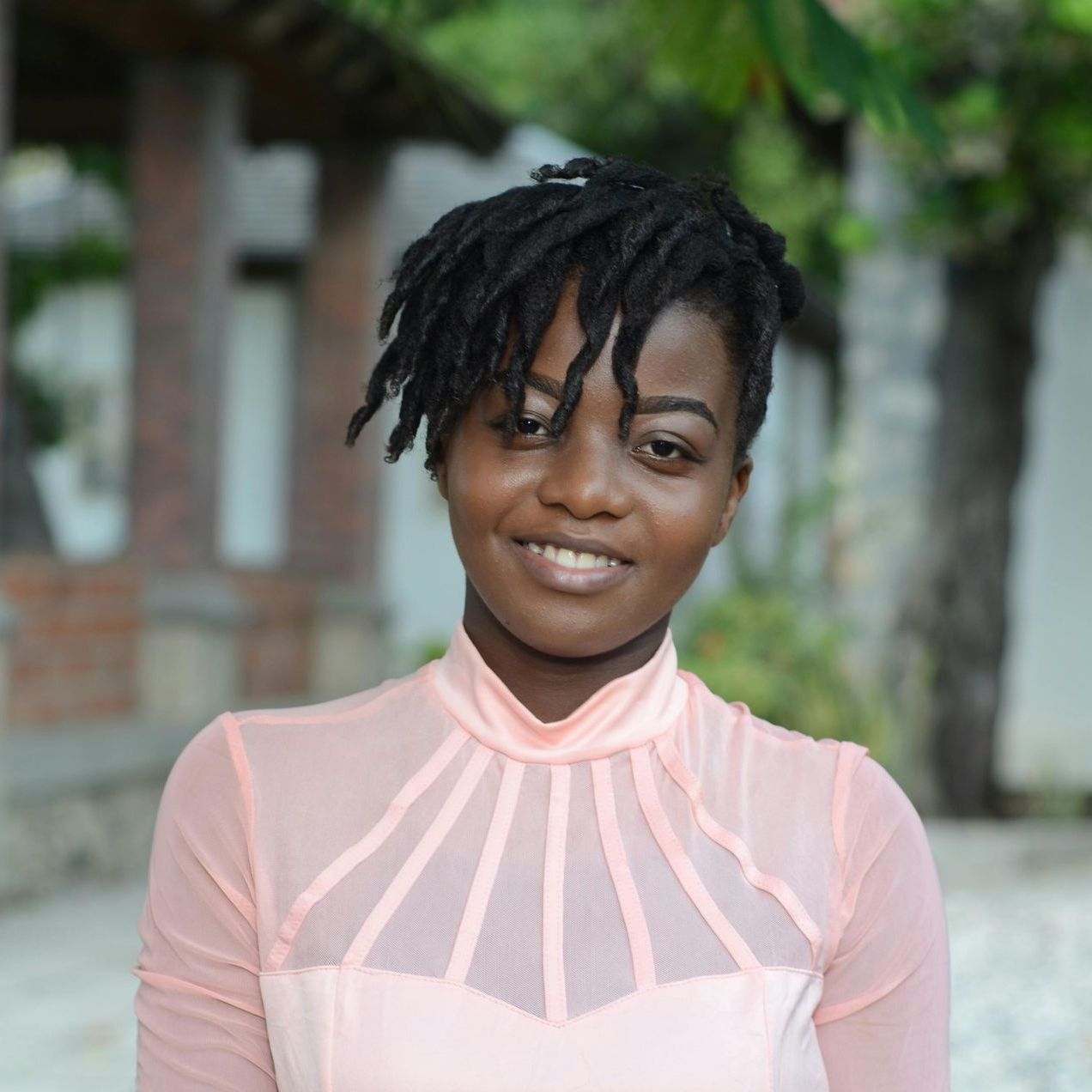 Une jeune femme noire sourit, vêtue d'une robe rose à décolleté haut, de dreadlocks foncés, à l'extérieur.