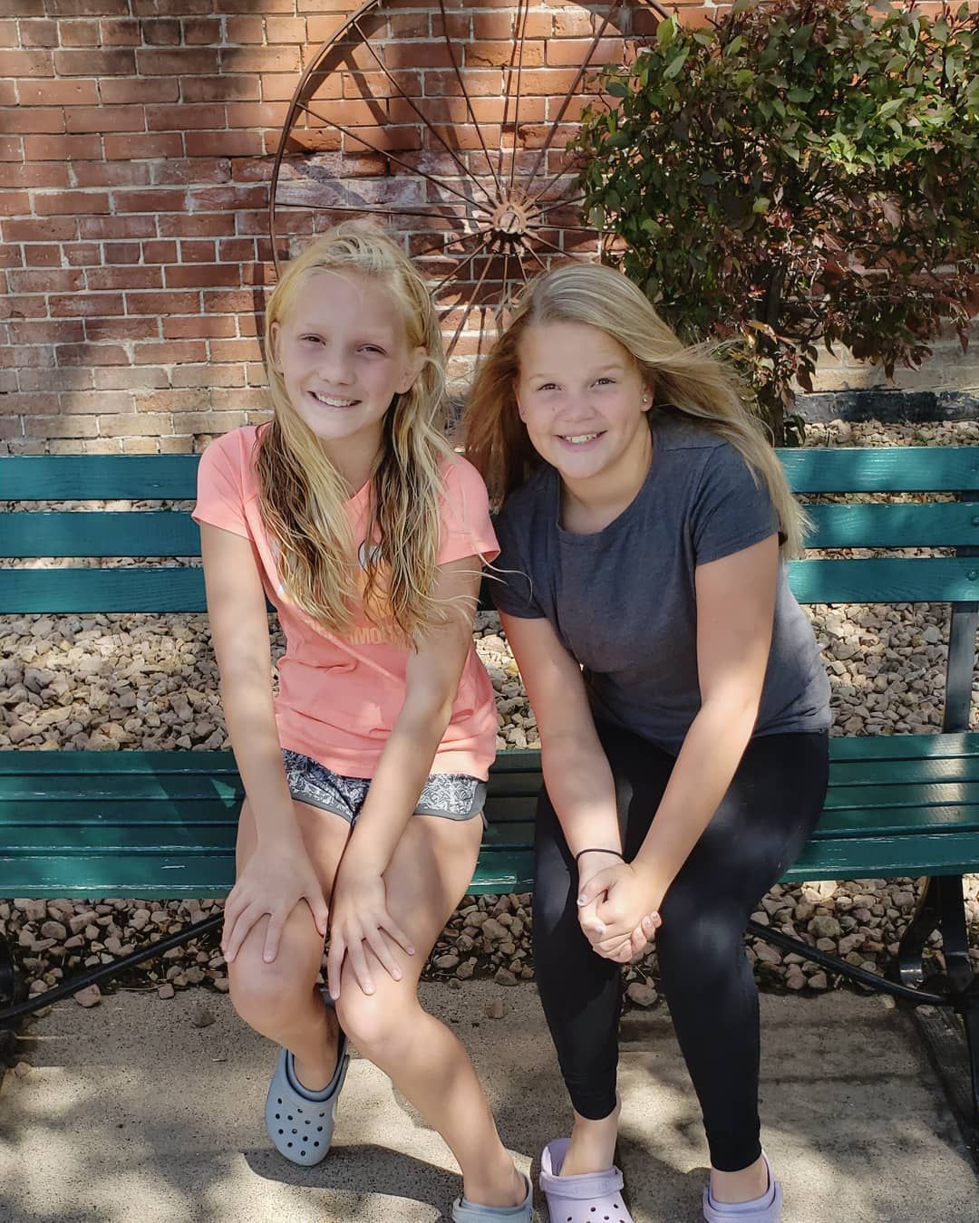 Two young girls are sitting on a green bench in front of a brick wall