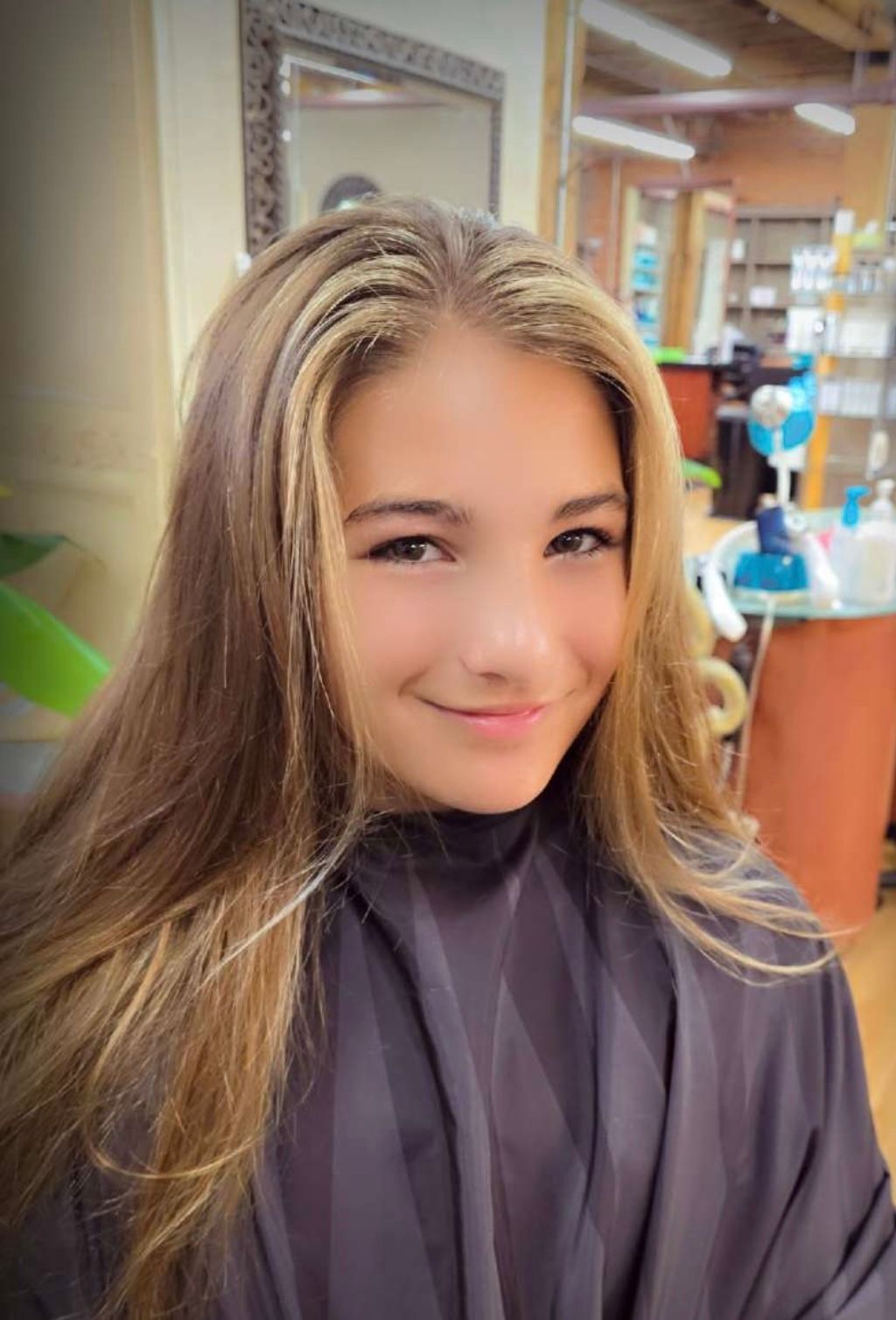 Young person with long blonde-highlighted hair smiles at the camera in a salon setting.