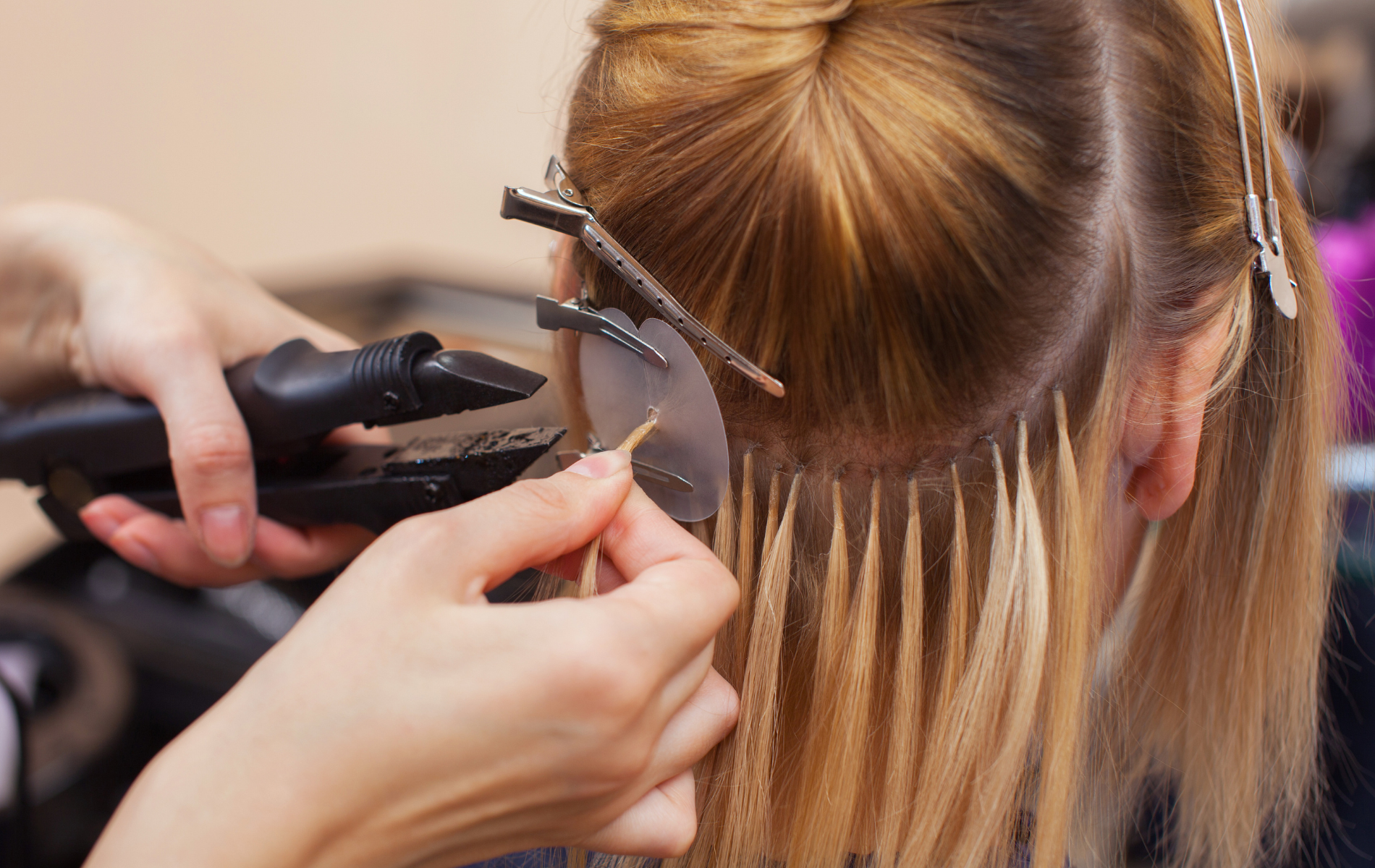 Hair extensions being applied to a woman's blonde hair by a stylist using a heat tool and applying bondings.