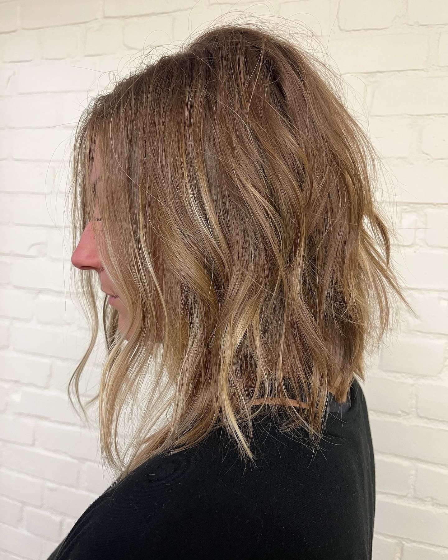 A woman with long blonde hair is wearing a black sweater and standing in front of a white brick wall.