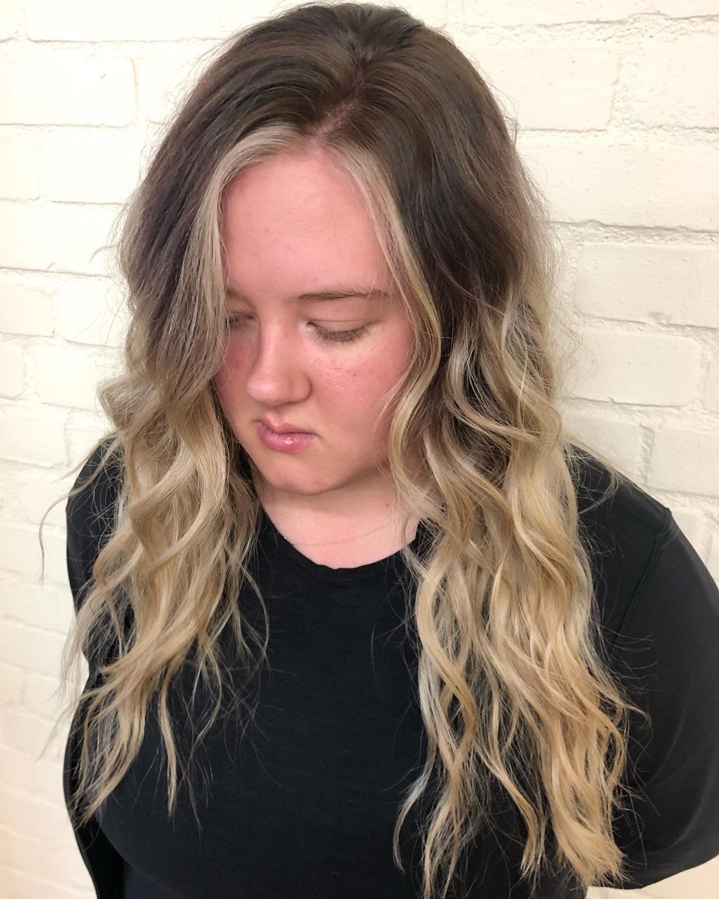 A woman with long blonde hair and a black shirt is standing in front of a white brick wall.
