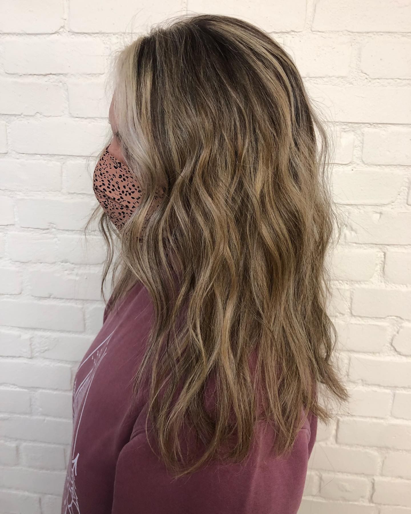 A woman wearing a mask and a purple sweater is standing in front of a white brick wall.