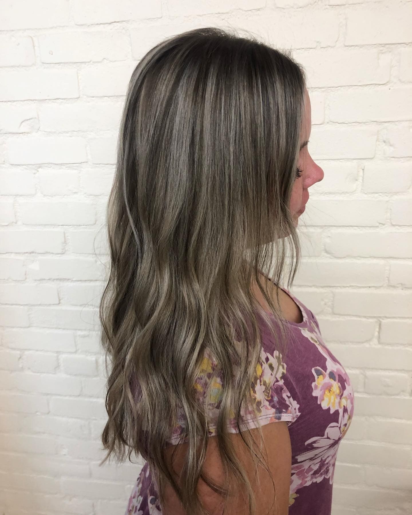 A woman in a purple dress with long hair is standing in front of a white brick wall.