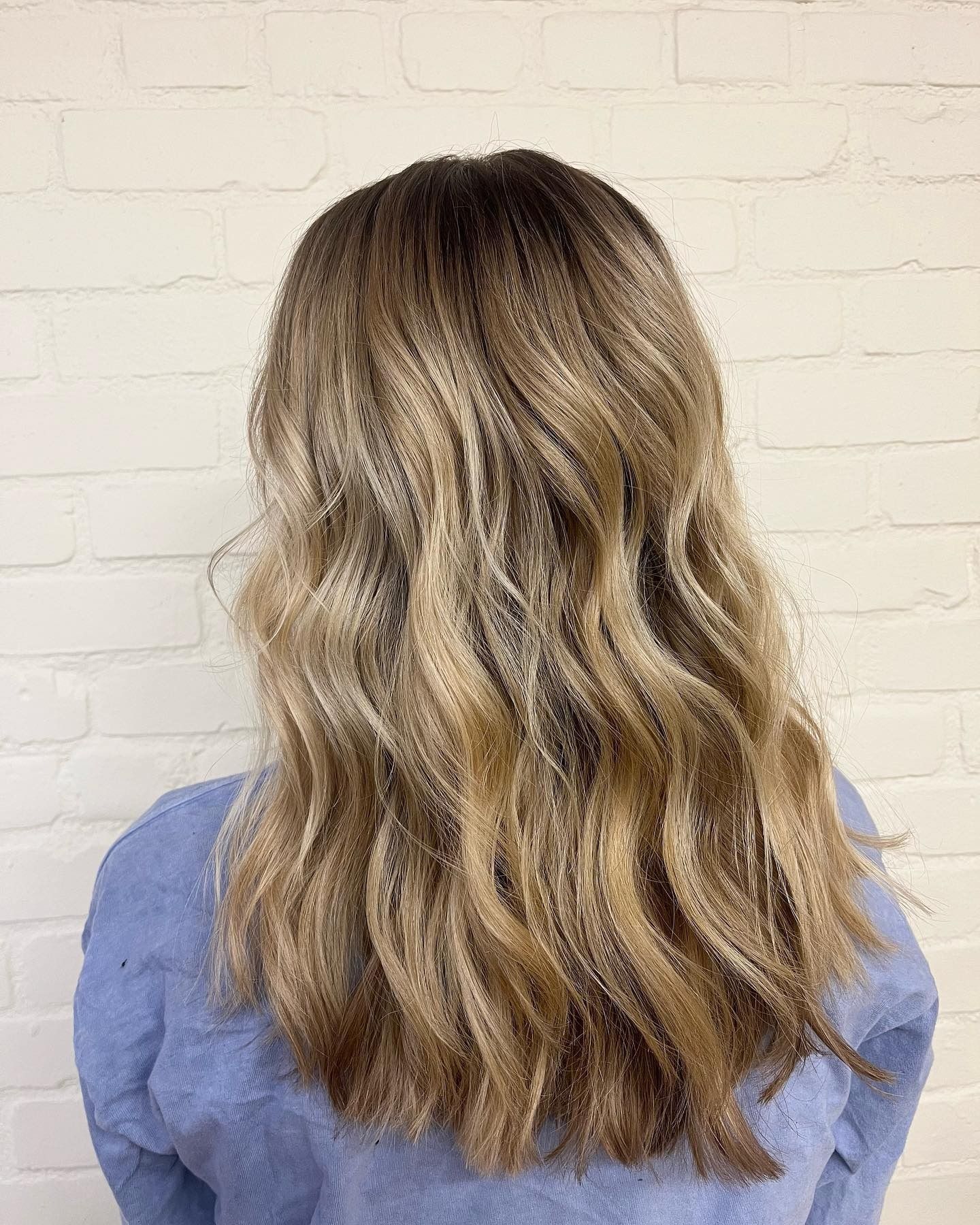 A woman with long blonde hair is wearing a blue sweater and standing in front of a white brick wall.