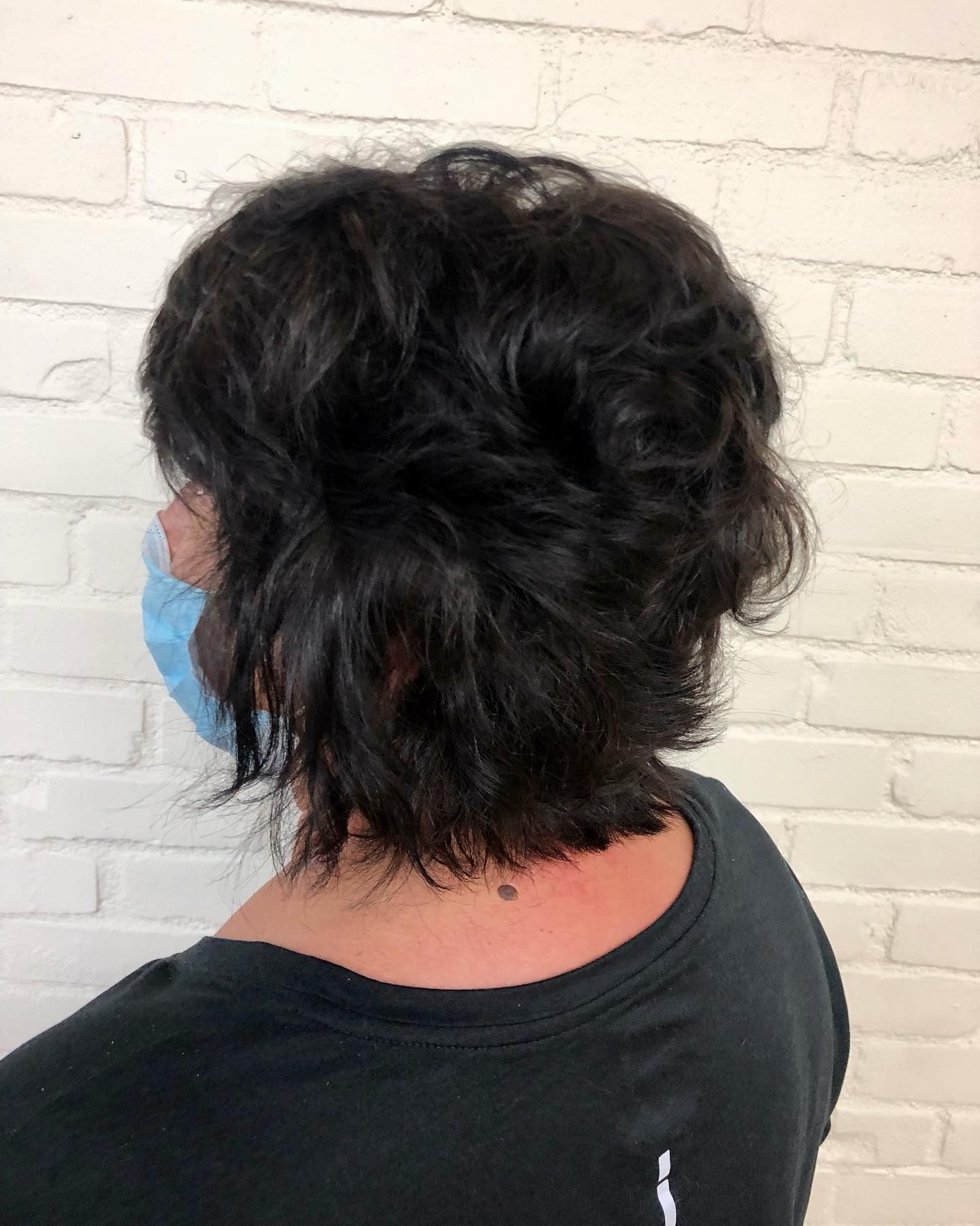 A woman wearing a mask and a black shirt is standing in front of a white brick wall.