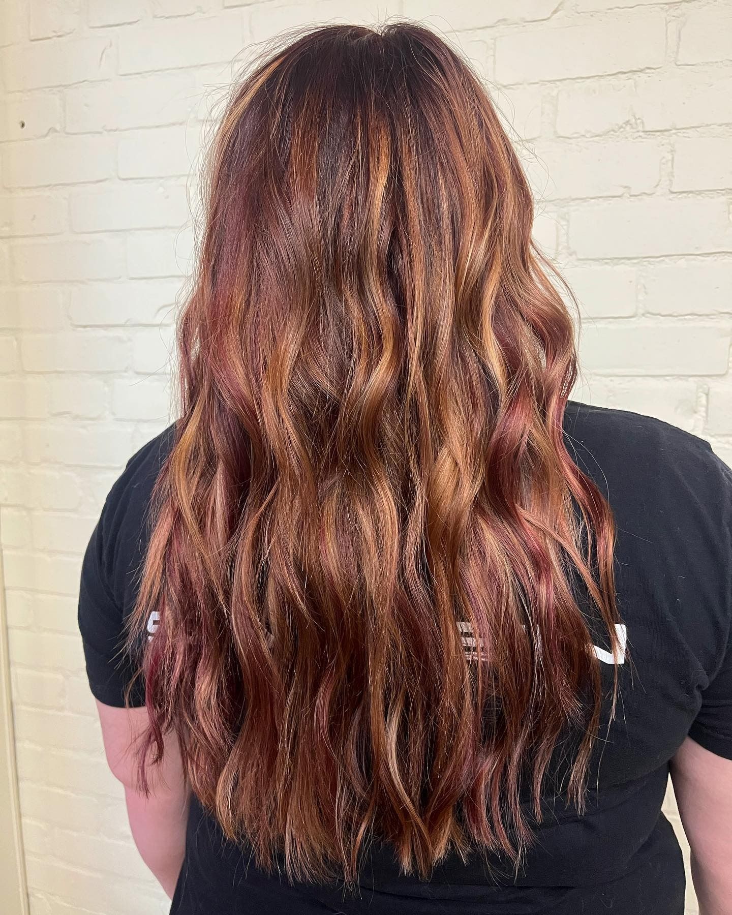 A woman with long red hair is wearing a black shirt and standing in front of a white brick wall.