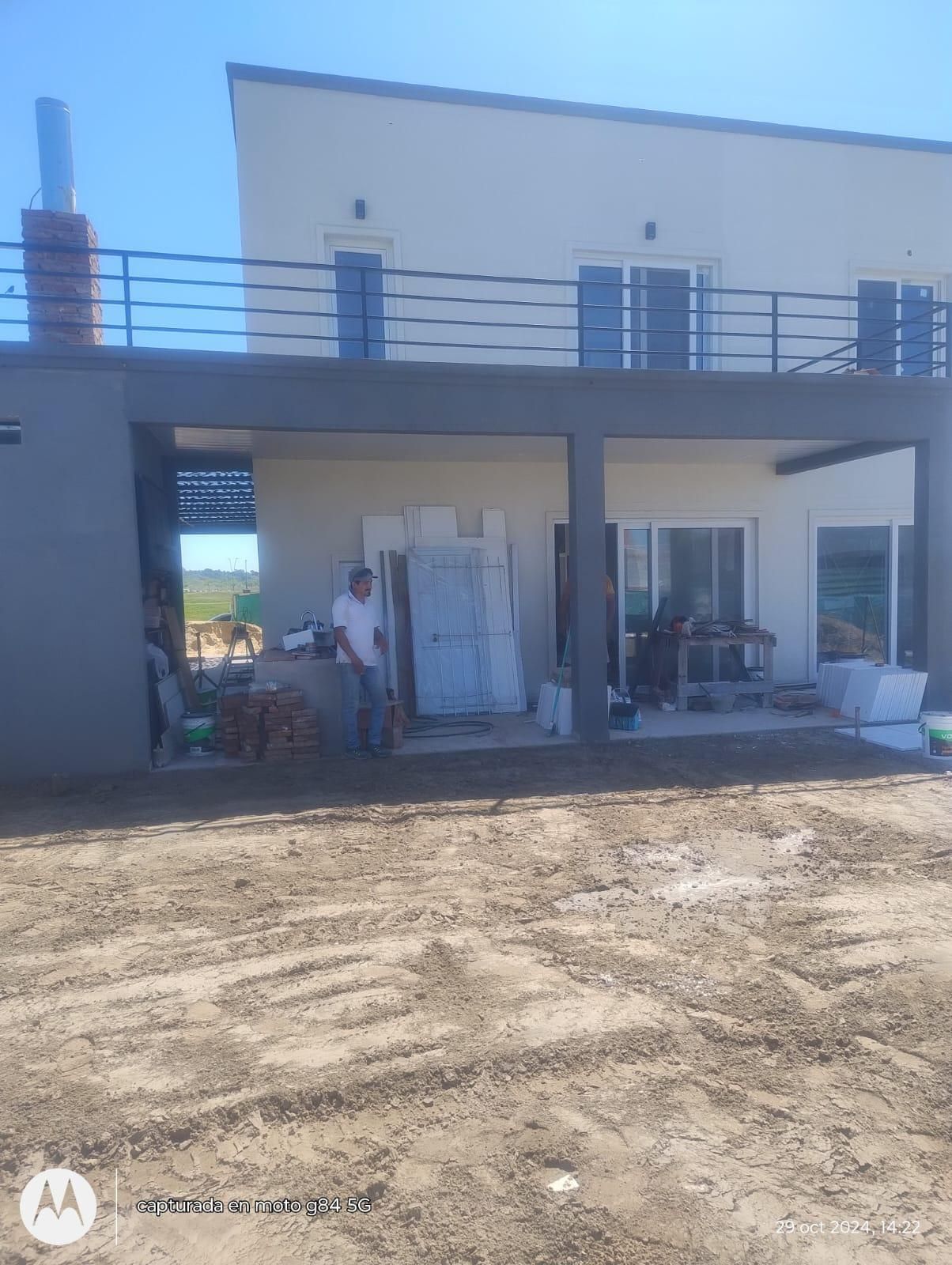 Hombre de pie cerca de una casa en construcción; colores neutros, entorno al aire libre.