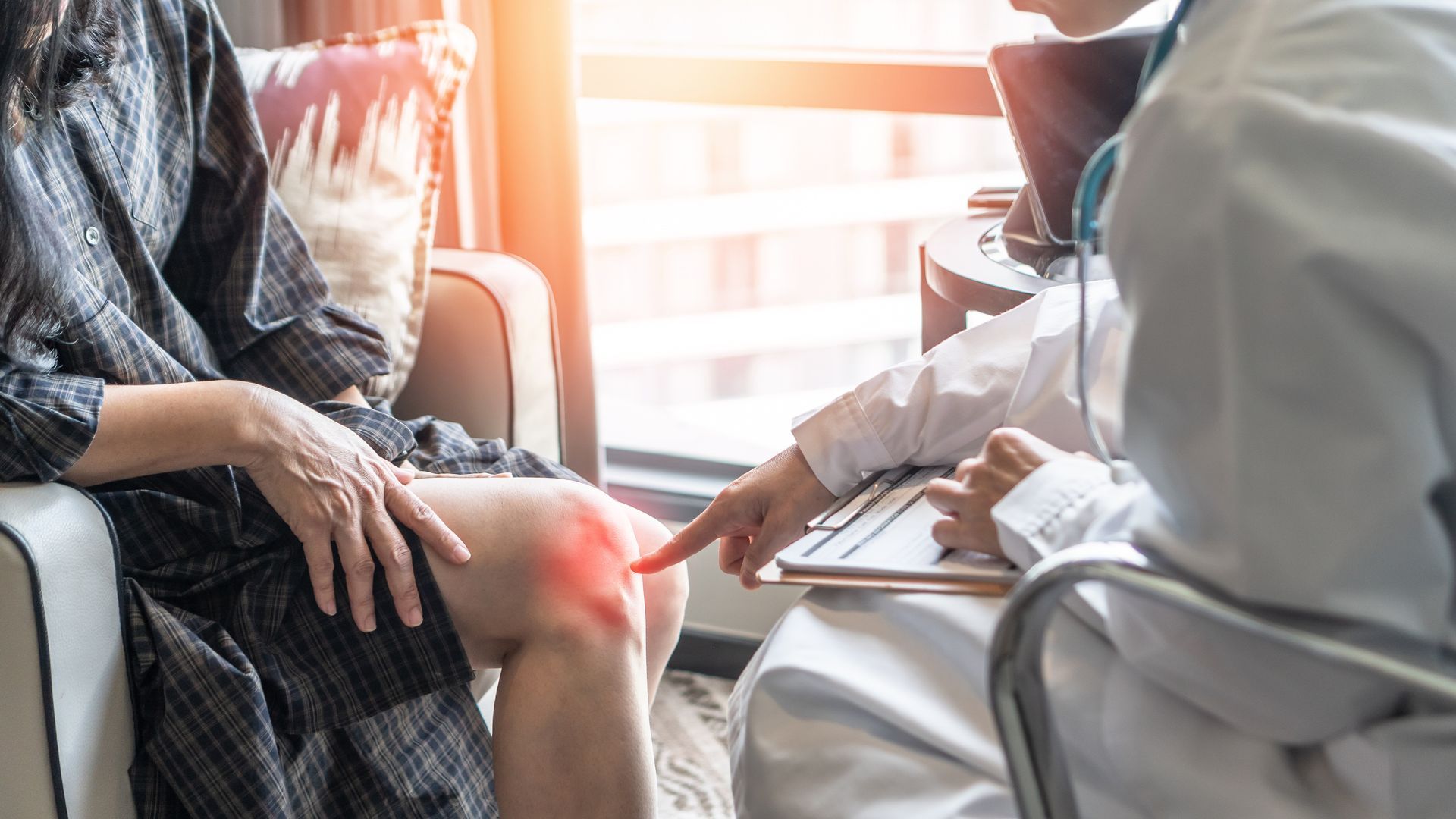 Der Arzt zeigt auf das rot markierte Knie des Patienten; die Konsultation findet in einer Arztpraxis statt.