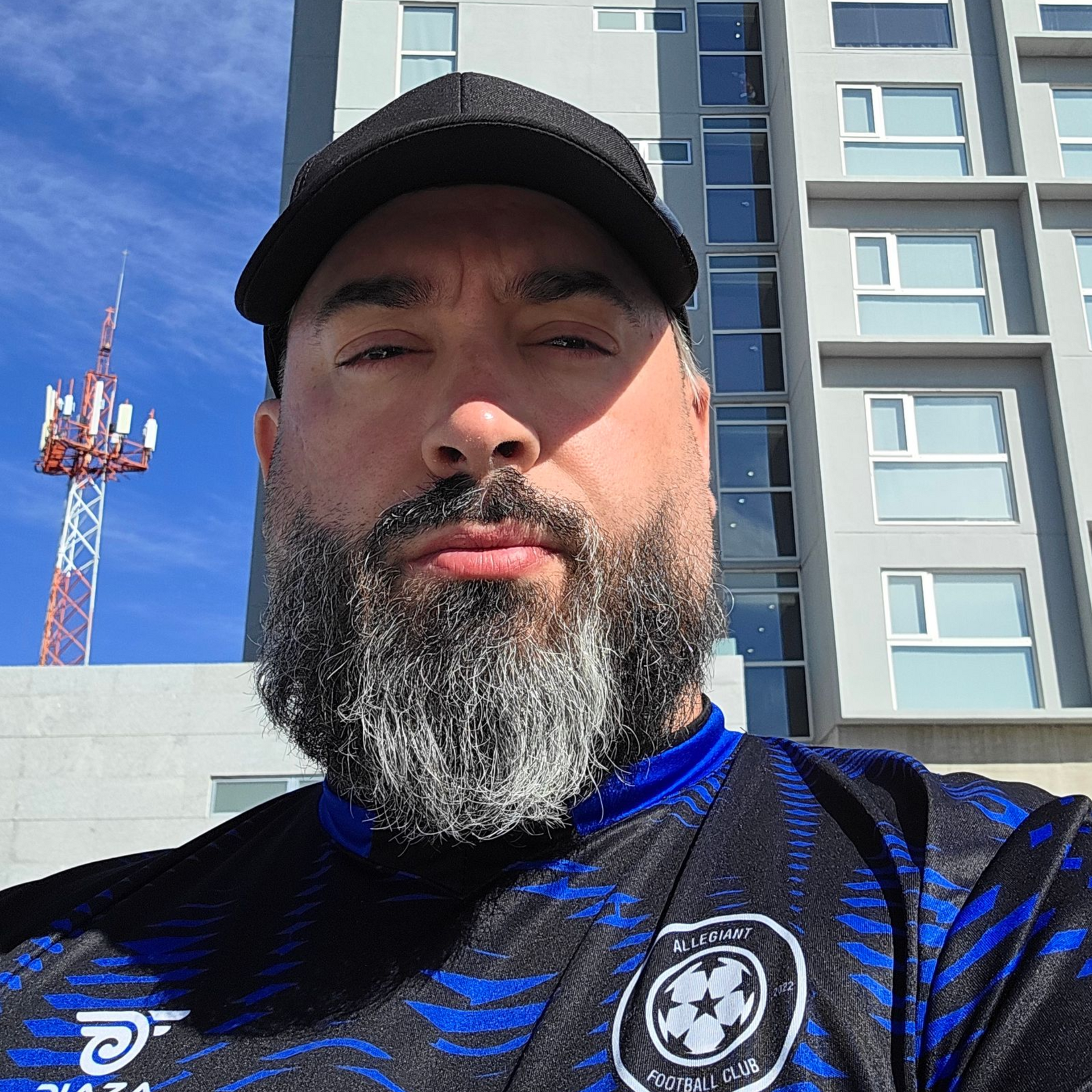 Man in black cap and gray beard, wearing a blue soccer jersey, in front of a building.