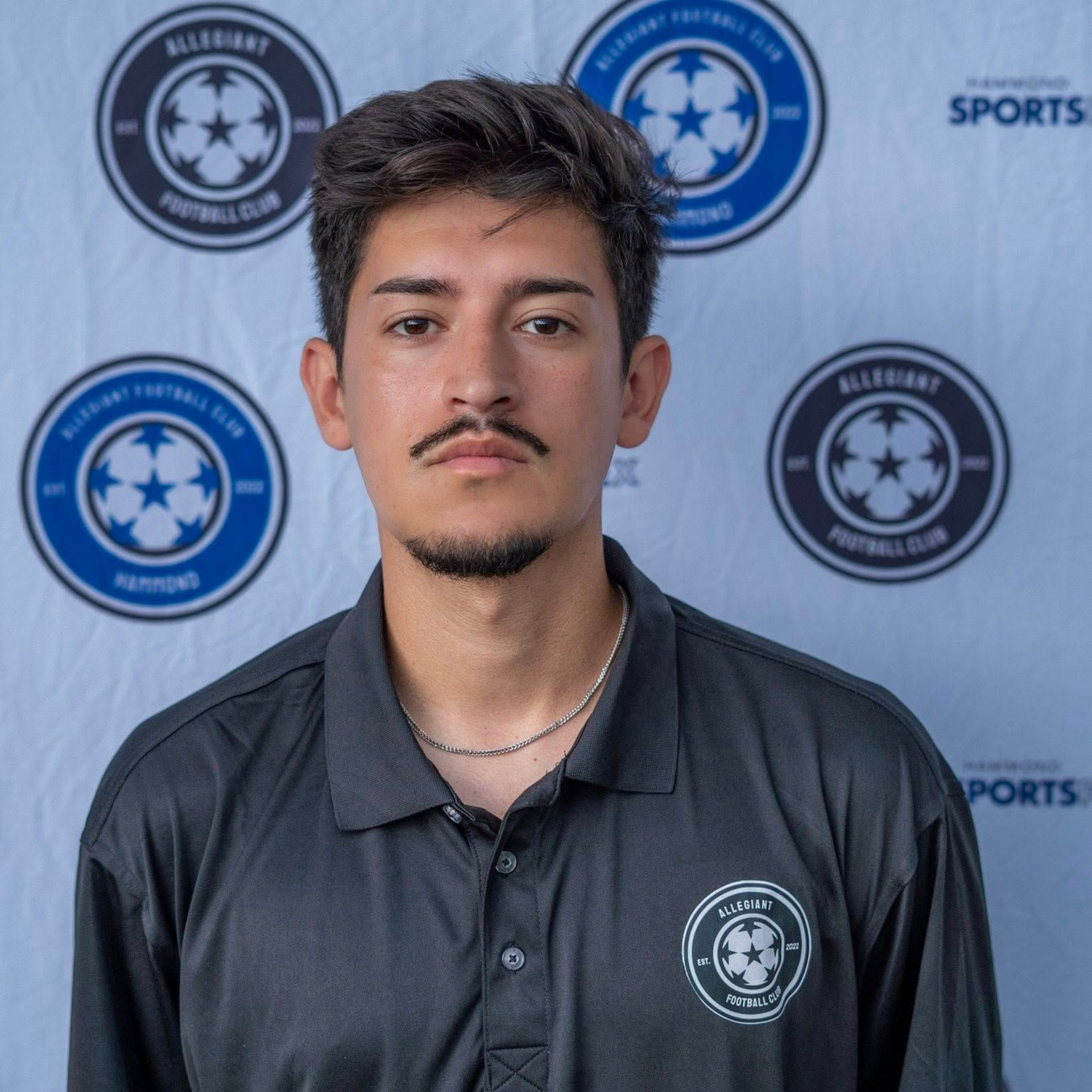 Man with dark hair and mustache in a black polo shirt, standing in front of soccer club banners.
