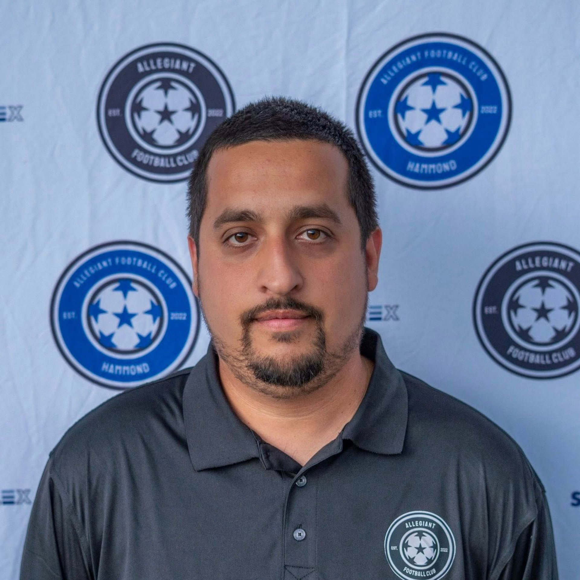Man in black shirt, posing in front of Allegiant Football Club logos.