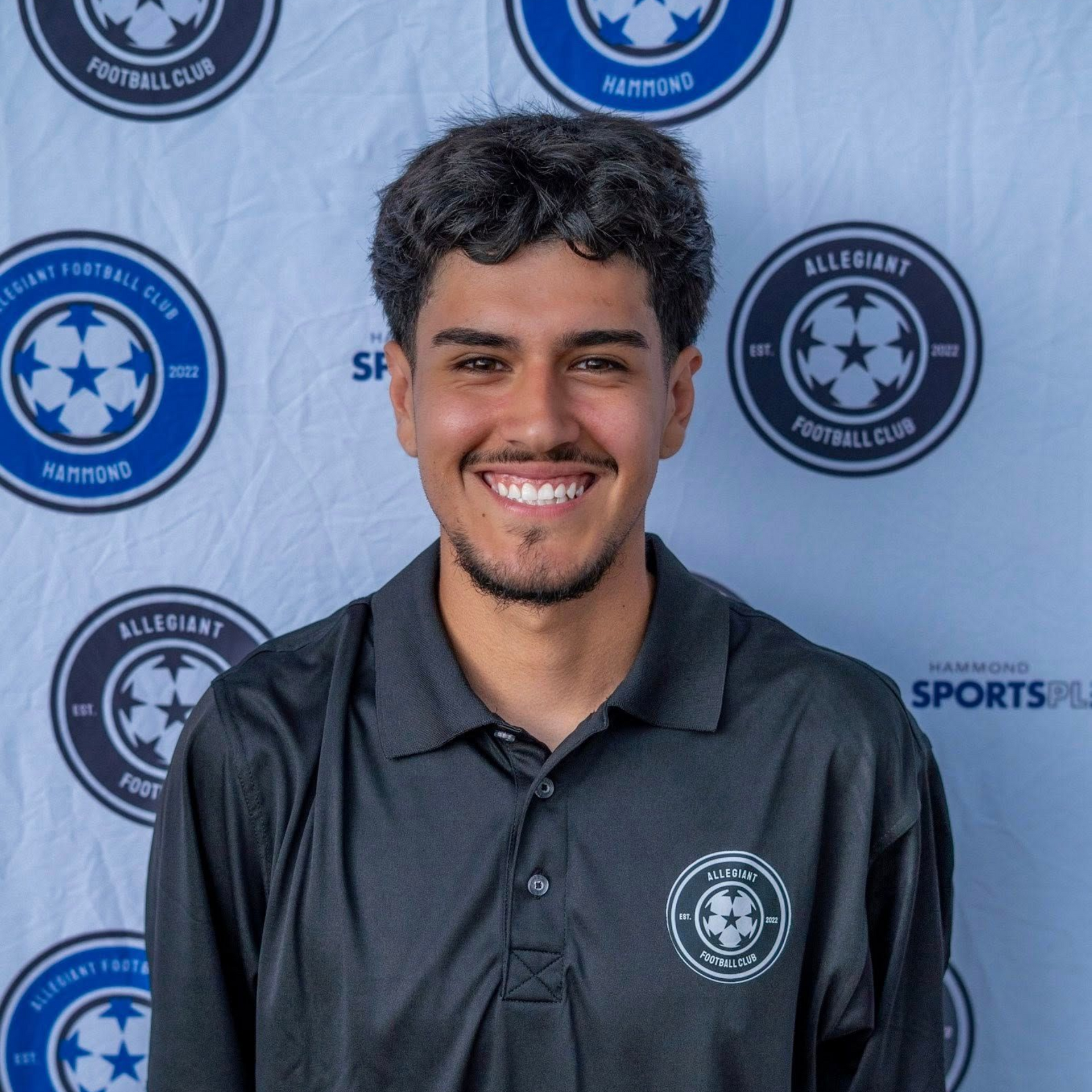Smiling man in black polo shirt with soccer club logo, standing in front of a backdrop with logos.