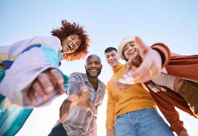 Group of four diverse friends smiling and reaching toward the camera from a low angle, set against a clear sky.