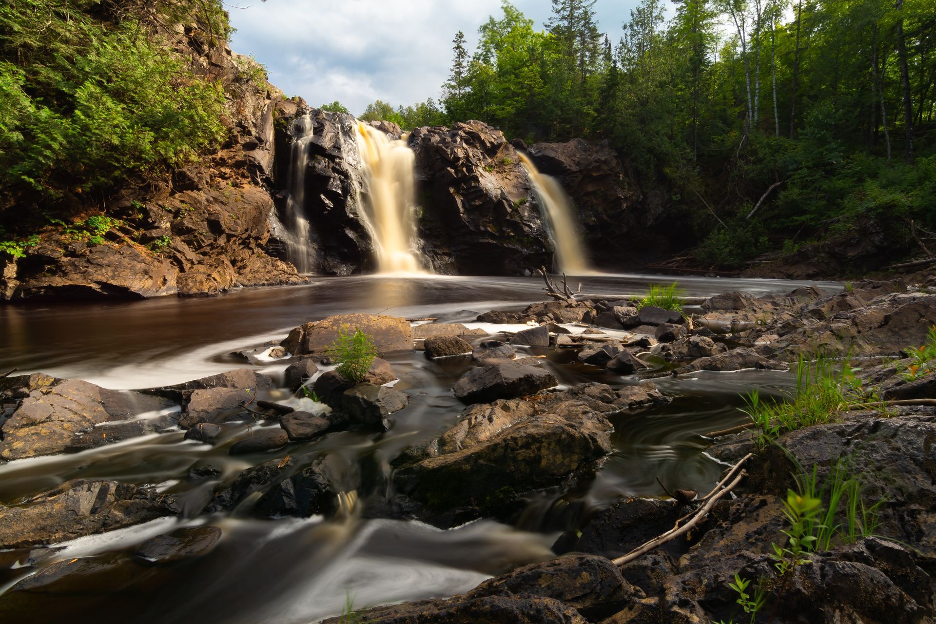 A long, empty road stretches toward mountains under a cloudy sky. Green hills and fields border the asphalt.
