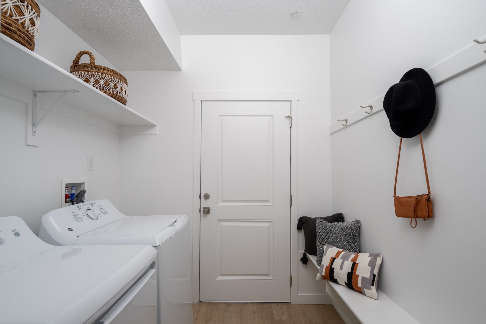 White laundry room with washer and dryer, open shelves, door, bench, and coat rack.