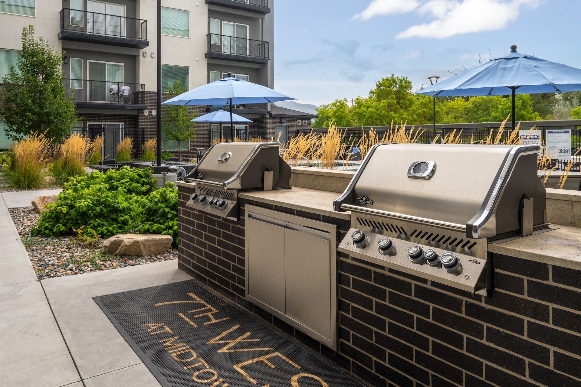 Two outdoor grills on a brick structure with a building in the background, sunny day.