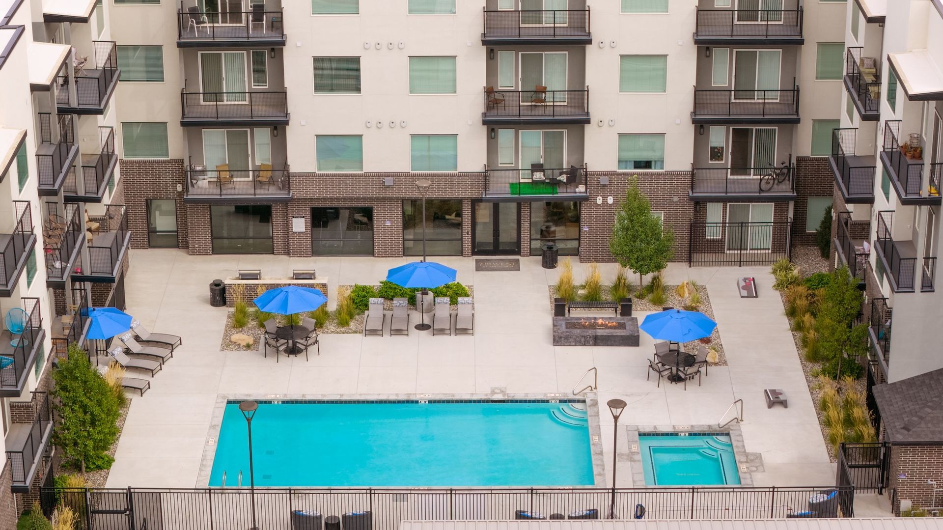 Apartment courtyard with pool, lounge chairs, tables, and blue umbrellas. White building with balconies surrounds.