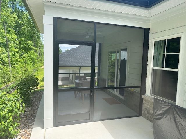A screened-in porch with a black door frame, white support columns, and adjacent white siding and window.
