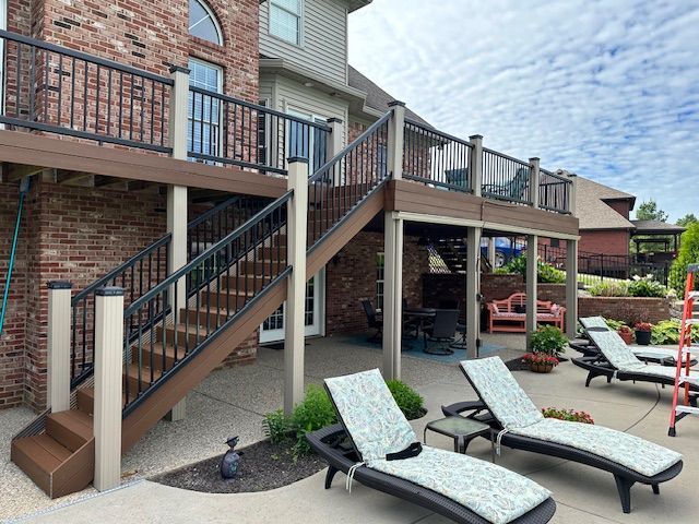 A multi-level composite deck with black railings and beige posts, featuring stairs leading to a concrete patio with loungers.
