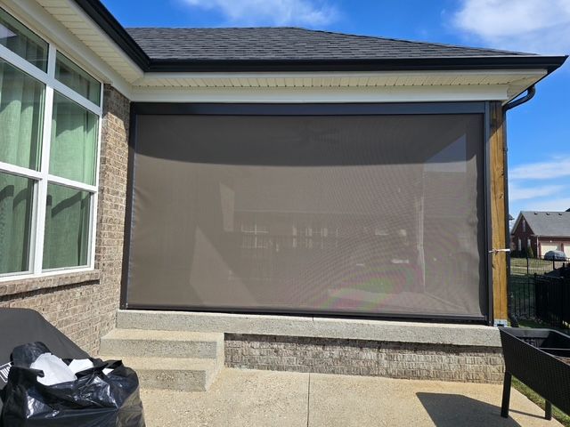 A brown retractable solar screen is installed on a brick home, enclosing a covered patio area above concrete steps.