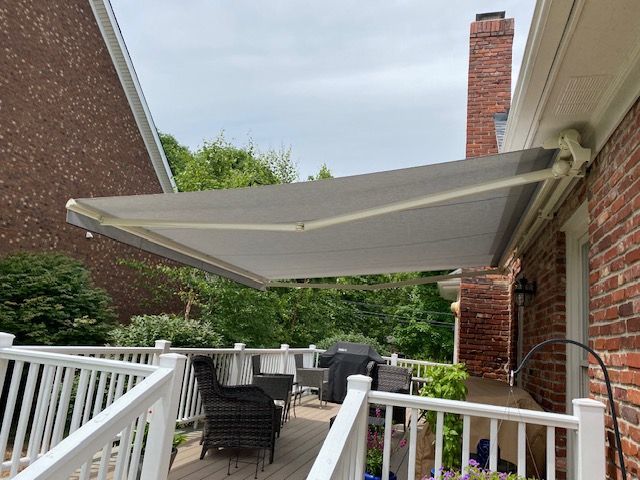 A retractable grey awning extends over a deck with patio furniture on the side of a brick house.