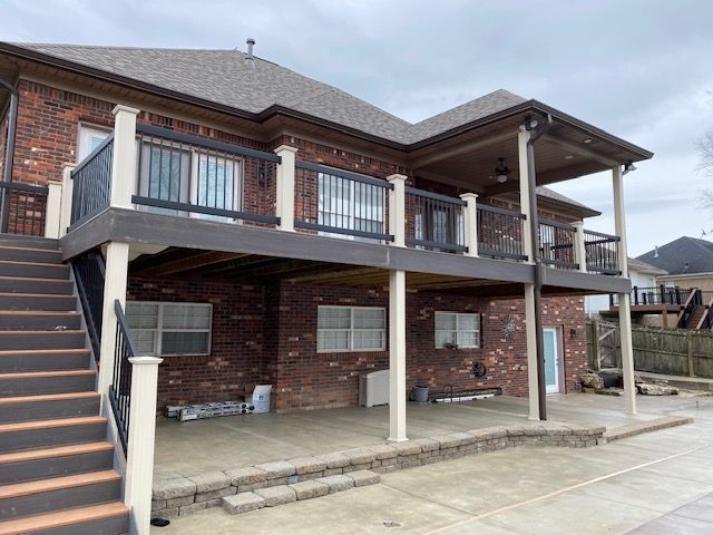 A two-story brick home featuring a raised wooden deck with railings, exterior stairs, and a concrete patio underneath.