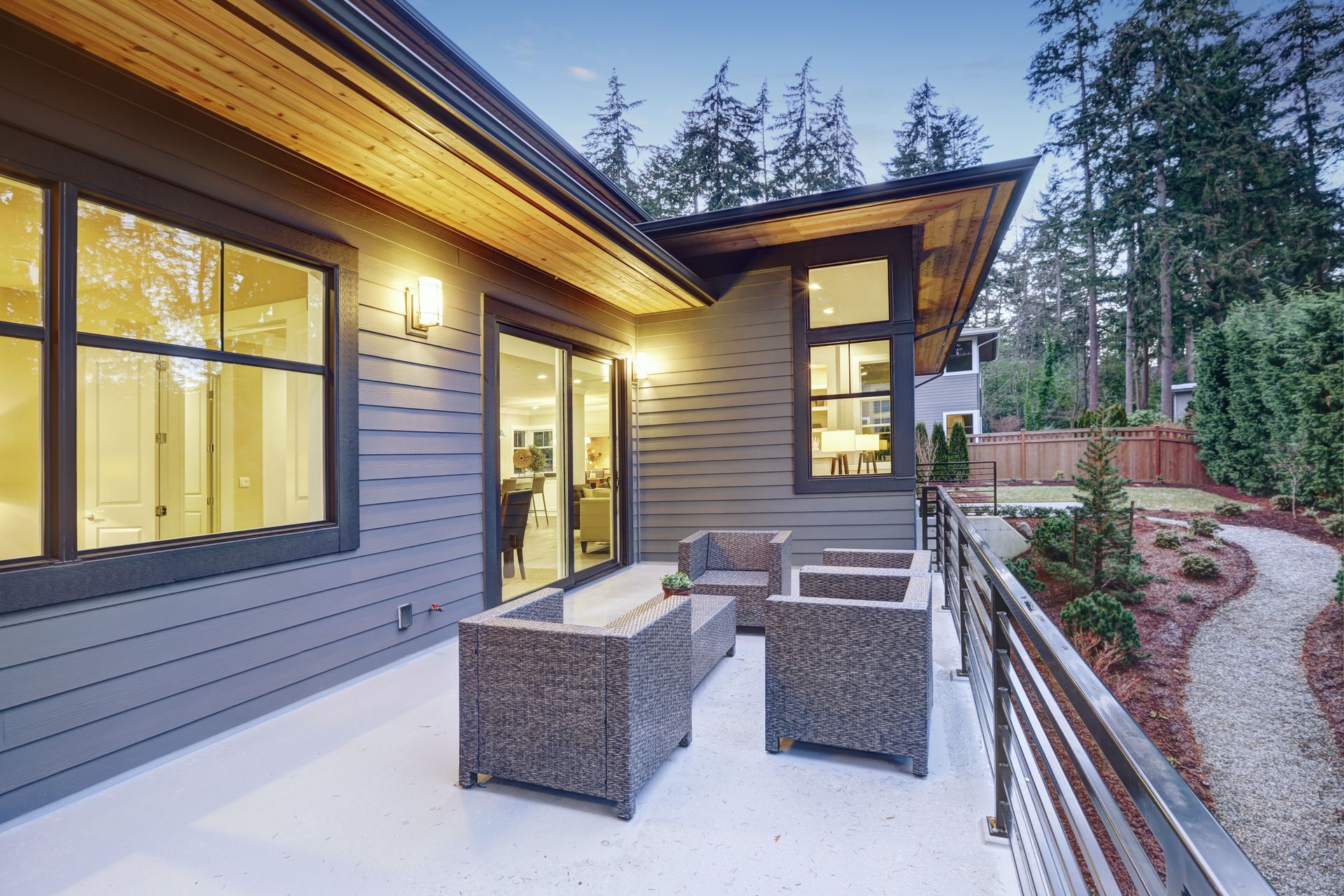 A contemporary gray patio with wicker furniture, a gravel path, and a wood-paneled ceiling at dusk.