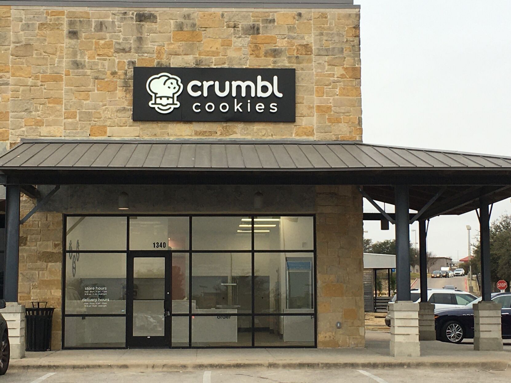 Crumbl Cookies storefront with large window, black sign, and awning. Building is light-colored stone.