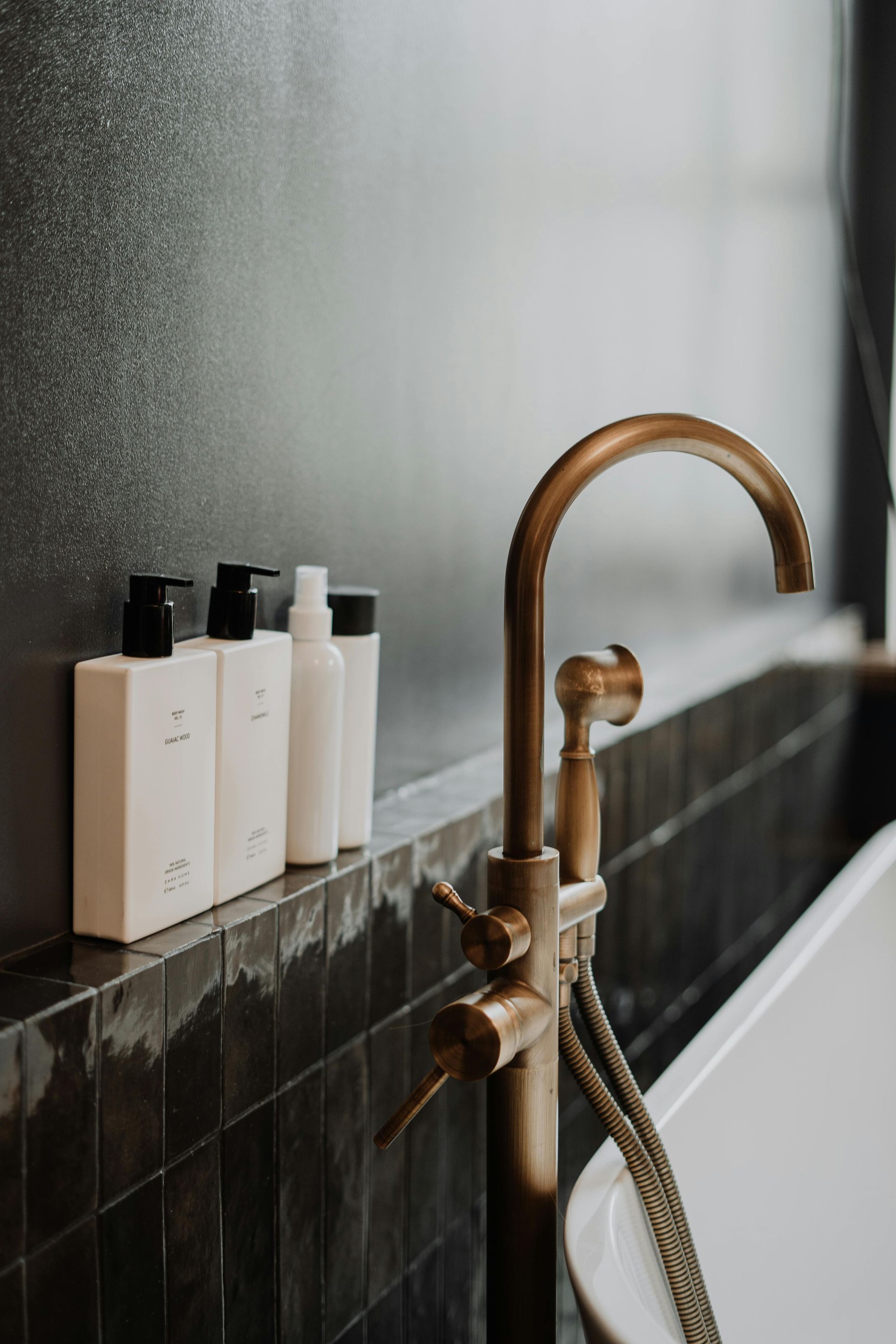 Bronze faucet with soap bottles on a tiled ledge against a dark wall.