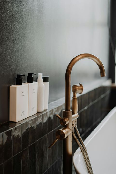 Bathroom with brass faucet and white toiletries on a dark tiled ledge against a dark wall.