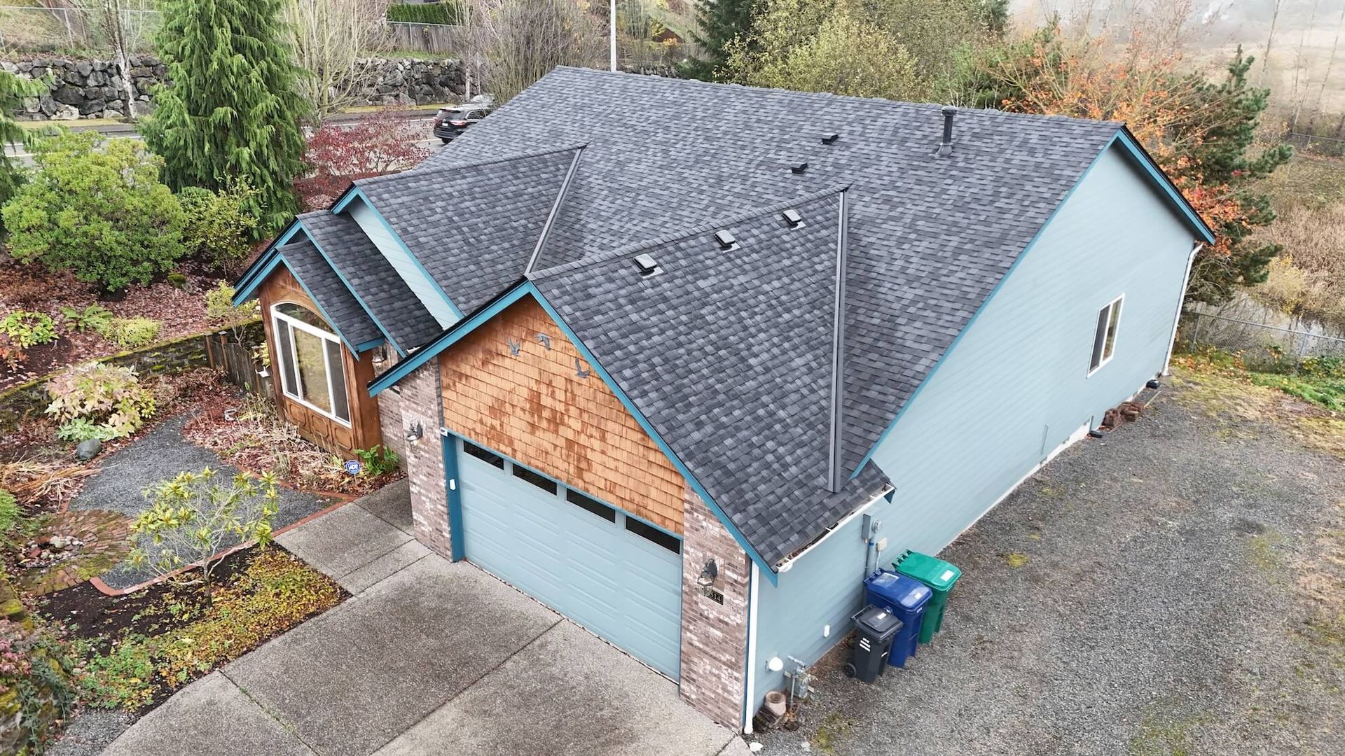 A light blue house with a gray shingled roof and brown brick accents. There's a garage, a driveway, and landscaping.