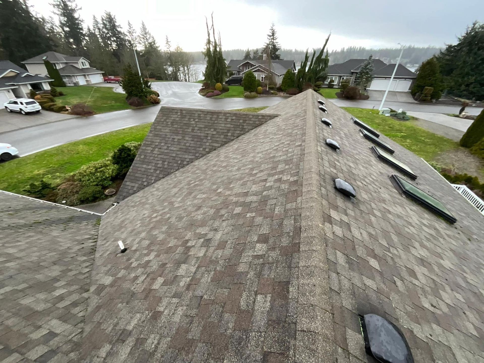 Overhead view of a brown shingled roof with several black vent caps; a residential street and green lawn are in the background.