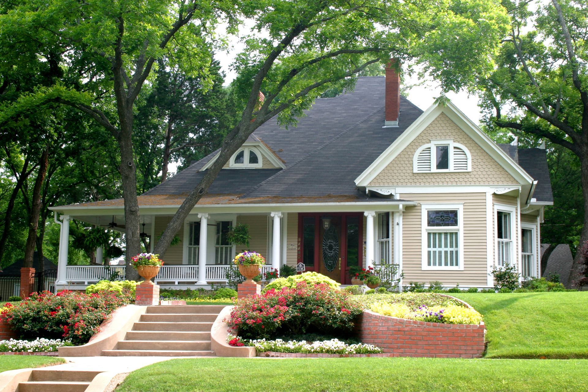 Tan two-story house with porch, set in a lush green yard with a brick staircase and flowerbeds.