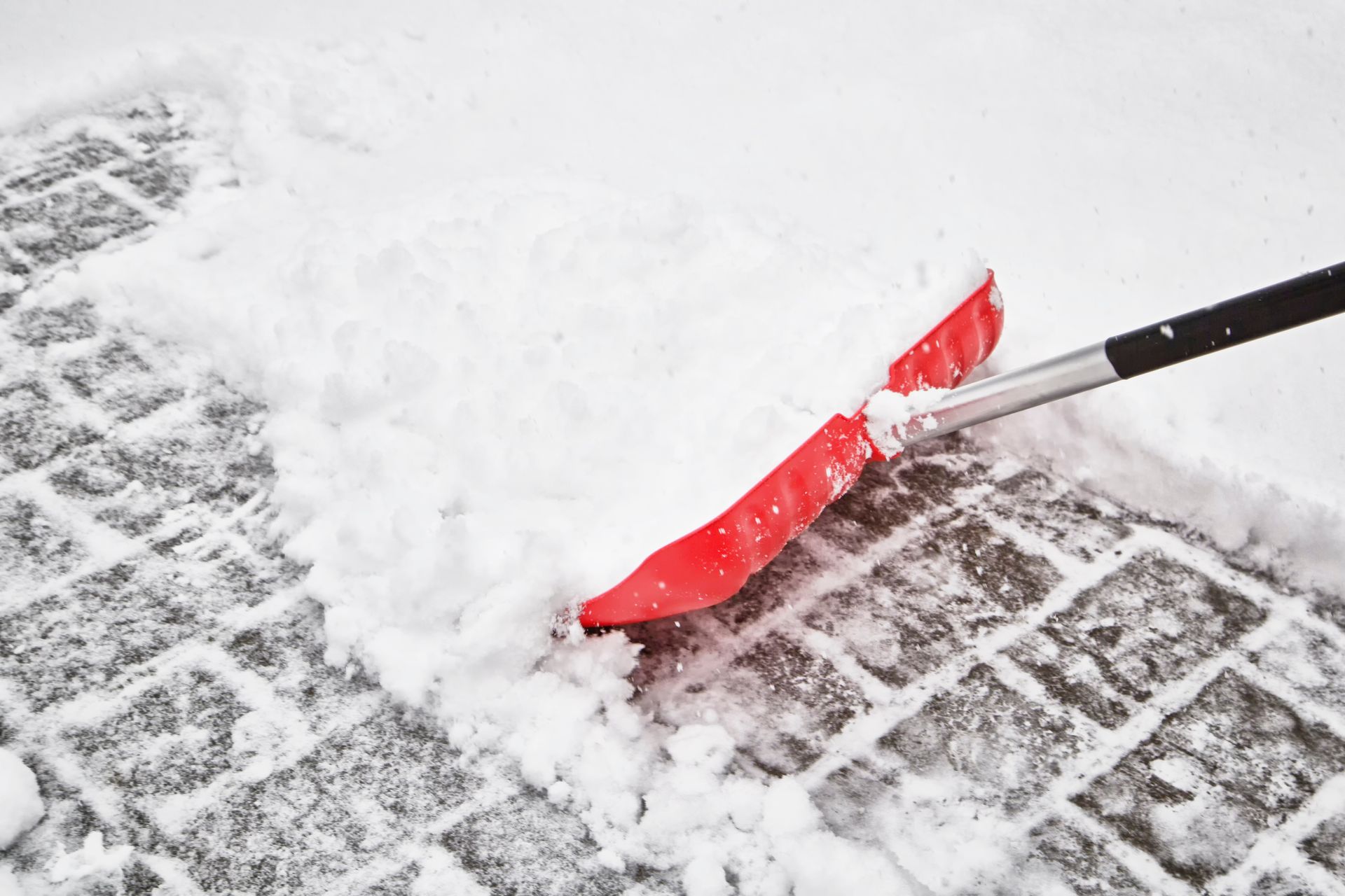 Red snow shovel clearing snow from a brick-patterned walkway.