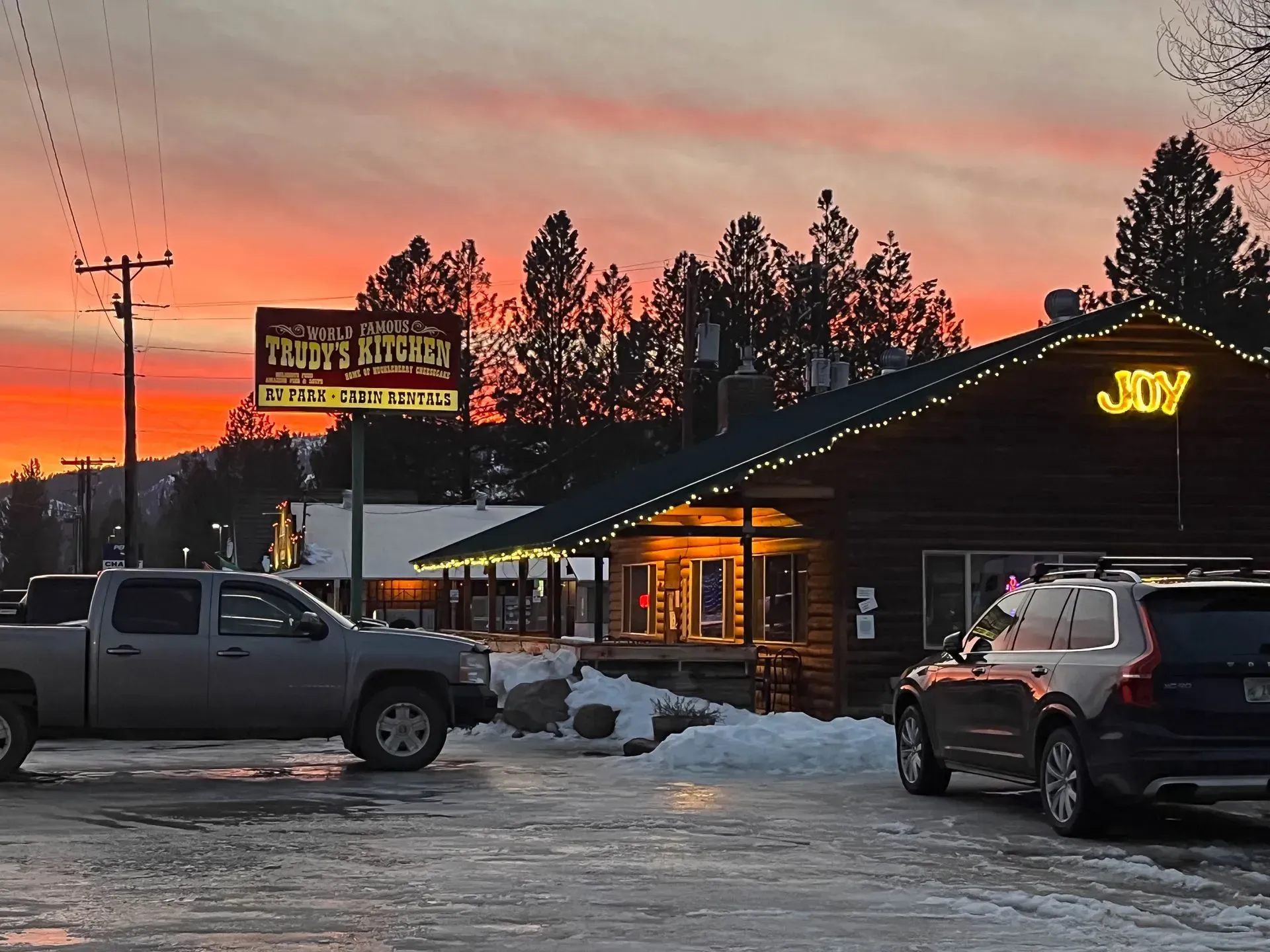 Restaurant at sunset; a log cabin-style building with a lit sign; cars and a truck parked outside.