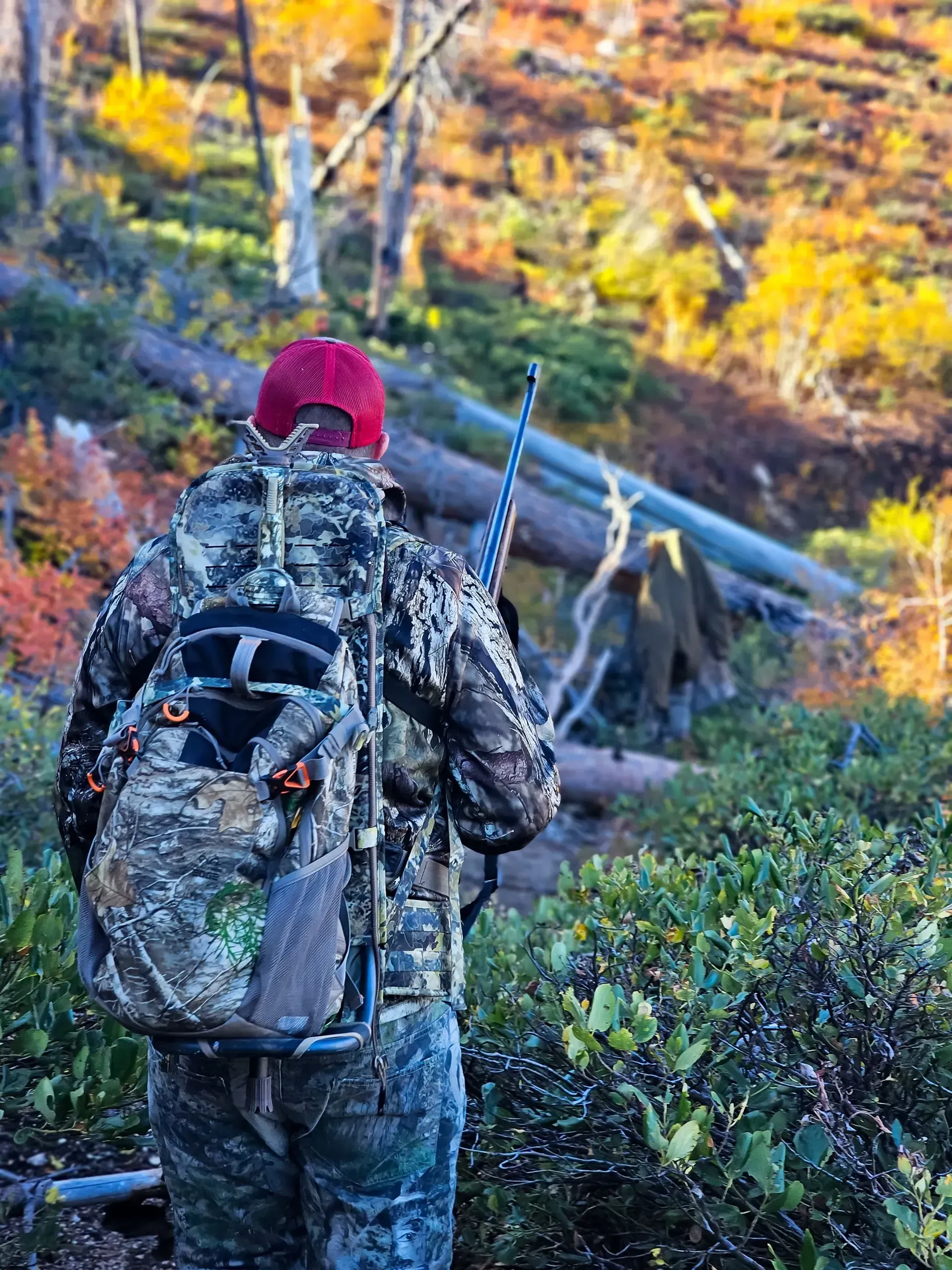 Hunter in camouflage gear with backpack, rifle, and red cap in a wooded area.
