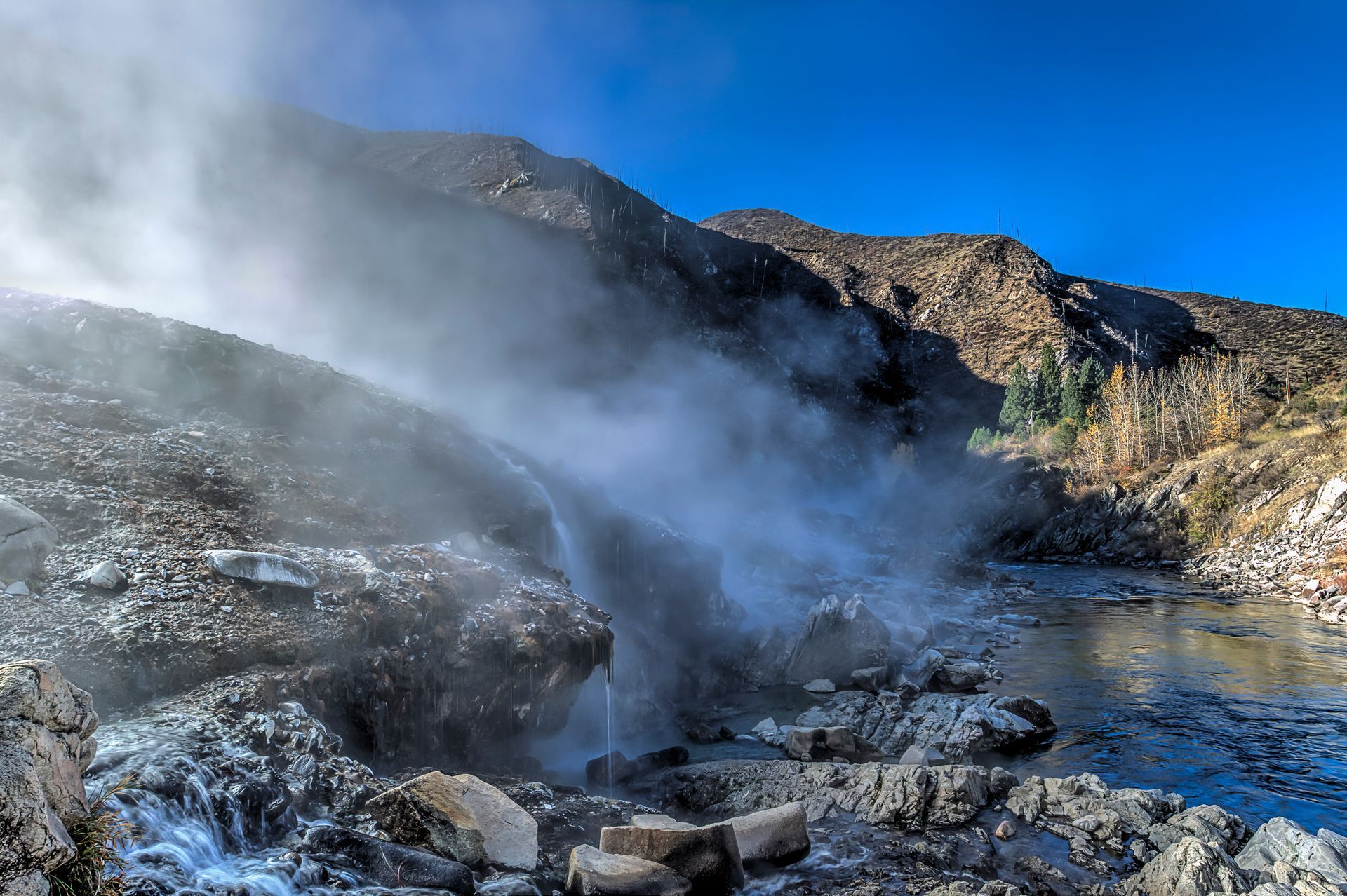 Steam rising from a rocky geothermal pool beneath a bright blue sky
