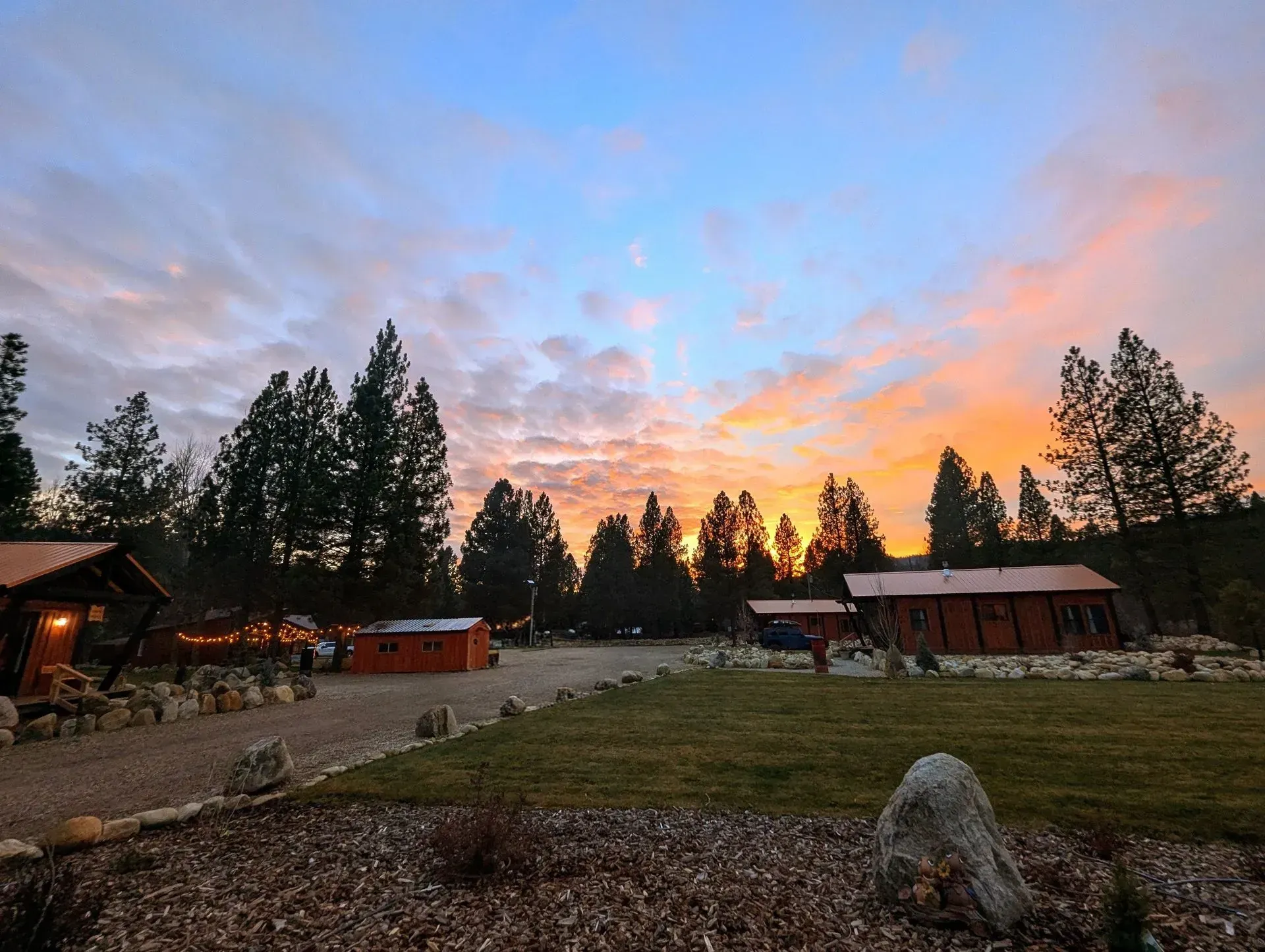 Sunset over cabins and pine trees. Orange and blue sky, gravel path, grassy area.